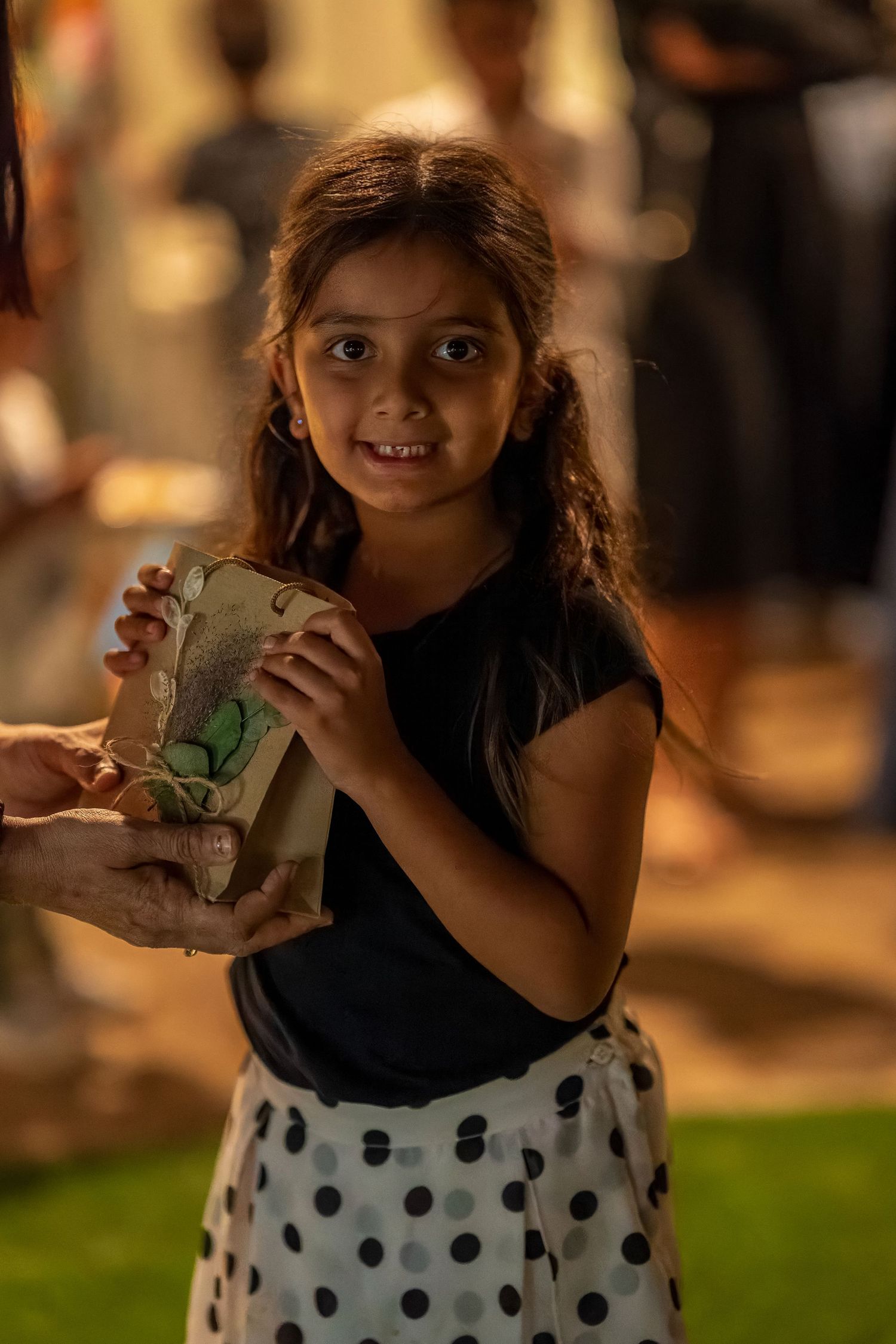 Child in polka dot skirt holds paper bag at outdoor evening event.