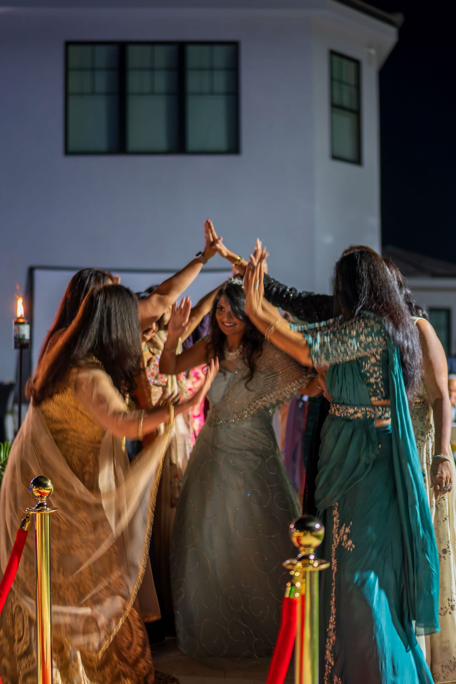A group of people in colorful formal attire raise their hands in celebration at an outdoor evening event.