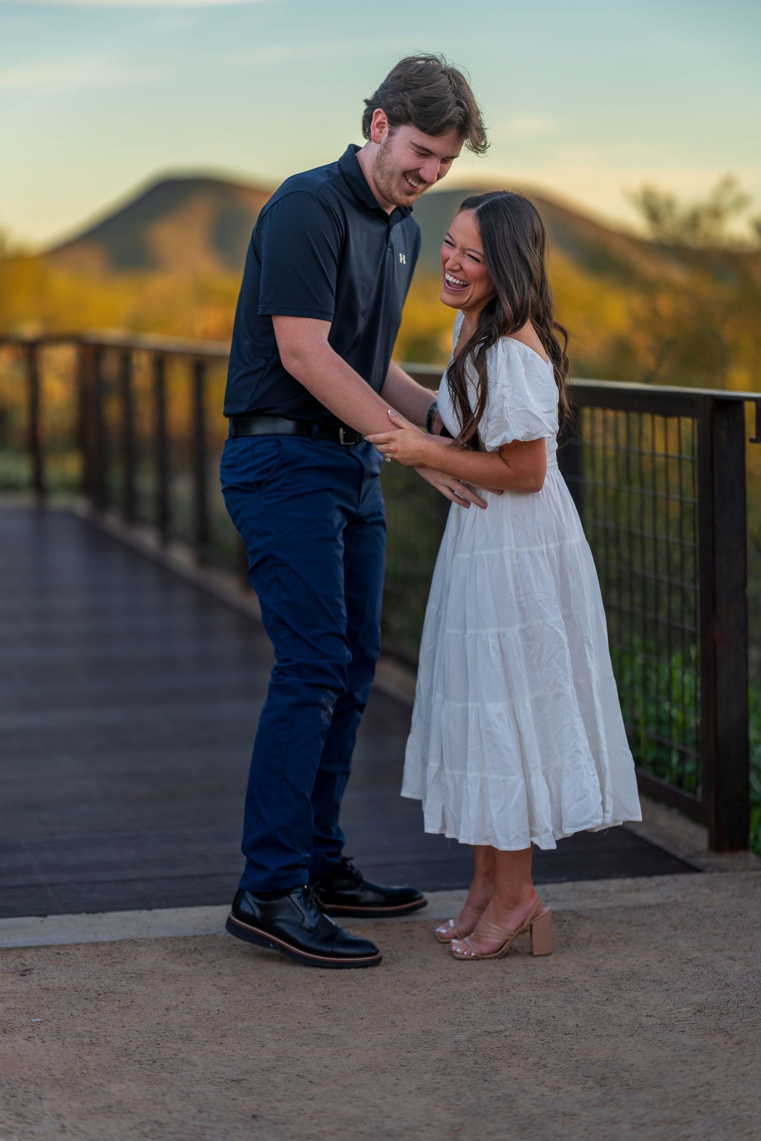 Two people embrace on a desert viewing platform with mountains and sunset in the background.