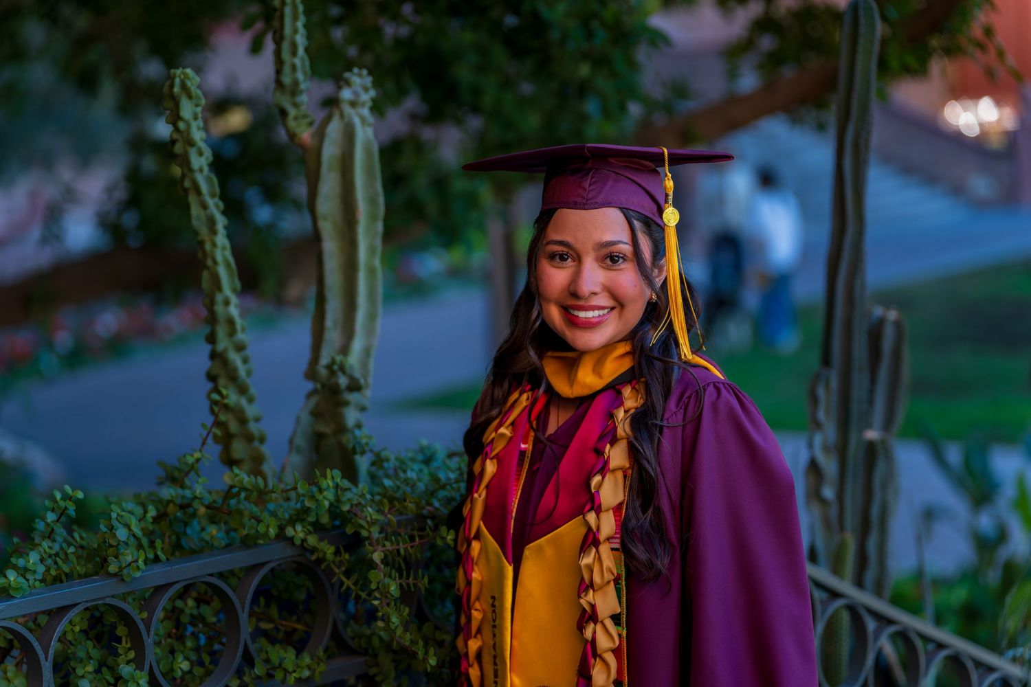 Graduate in maroon and gold academic regalia poses by decorative railing with cactus plants.