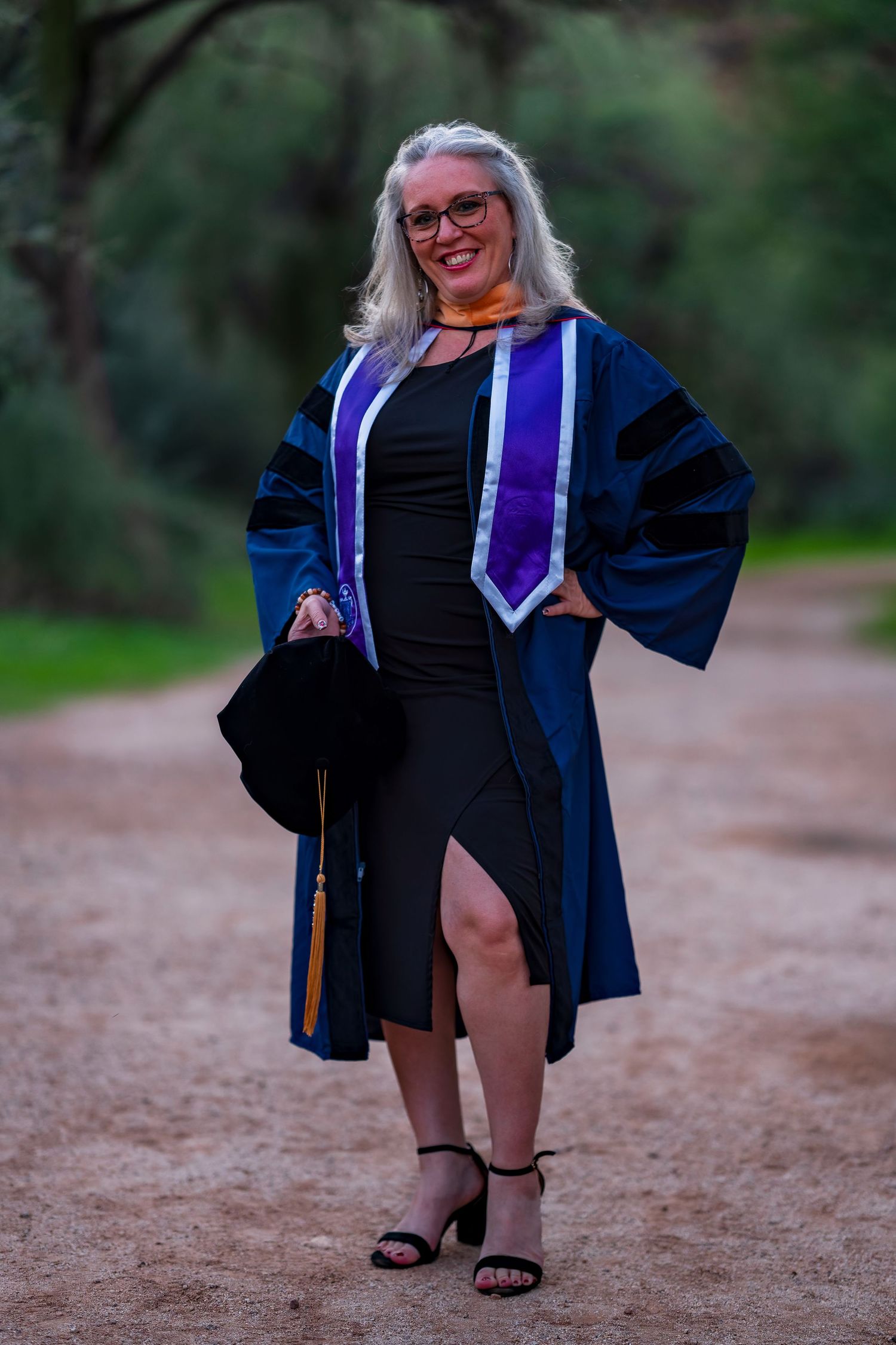 Graduate in doctoral regalia poses confidently on dirt path during sunset photo session.