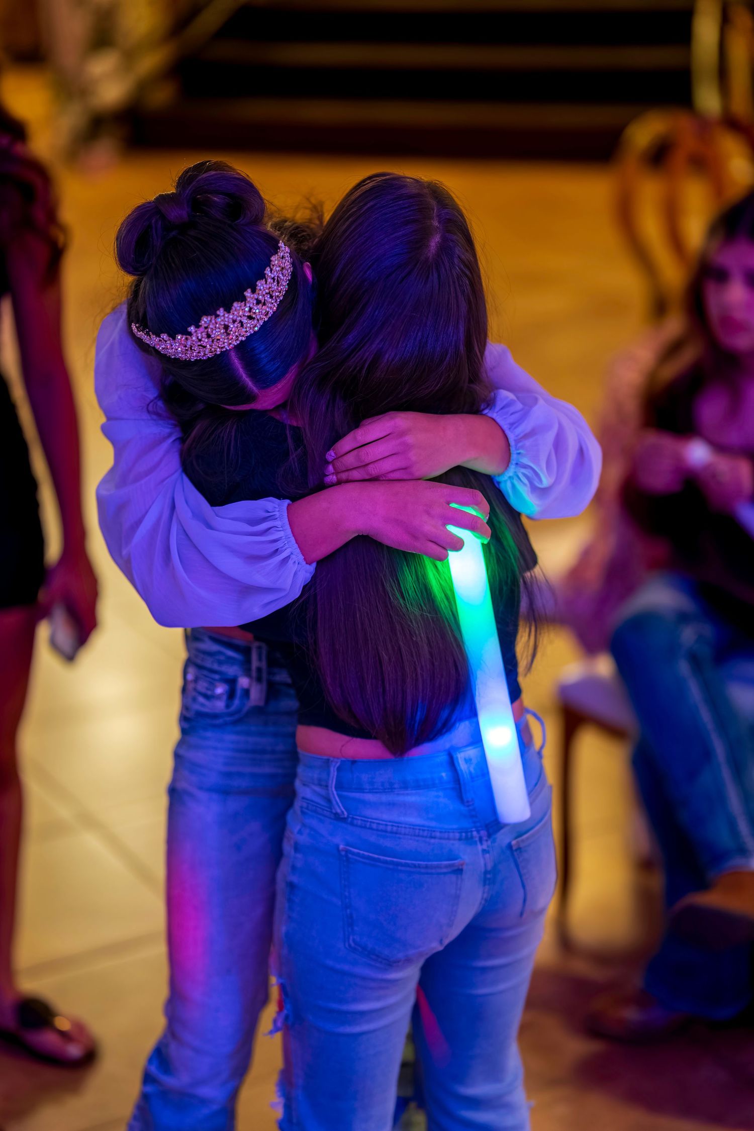 Two girls hugging at a party, one wearing a tiara and holding a glowing LED foam stick under colorful neon lights.