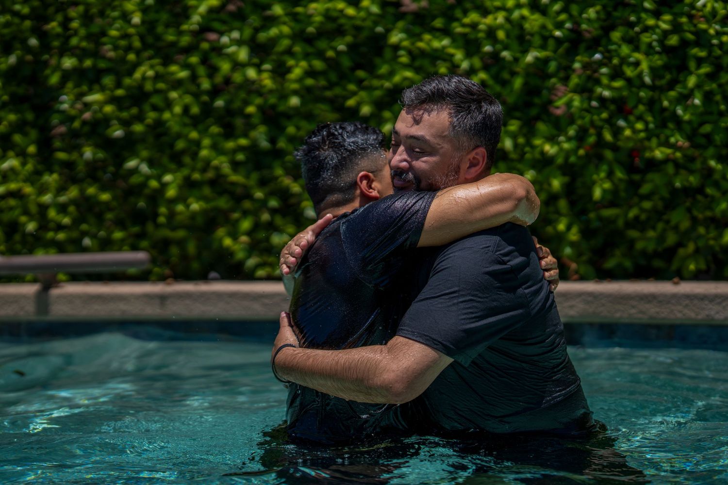 Two people sharing a warm embrace while standing in a swimming pool, surrounded by lush green hedges.