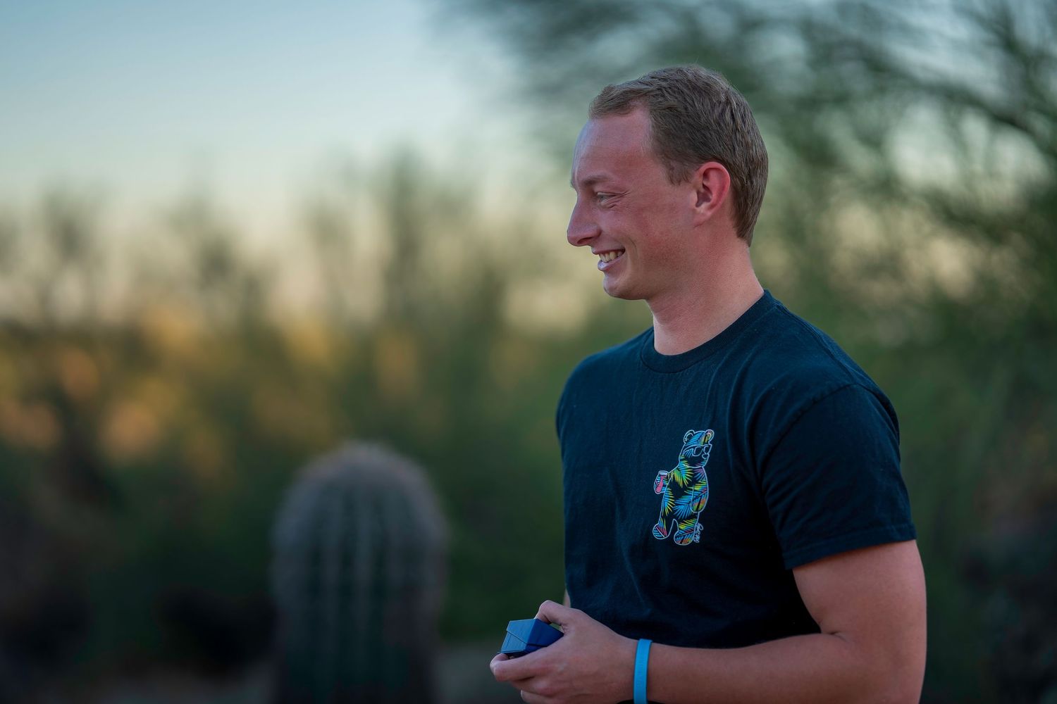 Smiling young man in black graphic t-shirt standing outdoors near cactus at dusk in desert landscape.