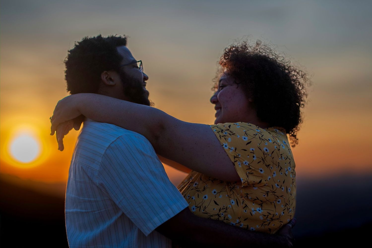 Couple embracing romantically against a vibrant orange sunset sky outdoors.
