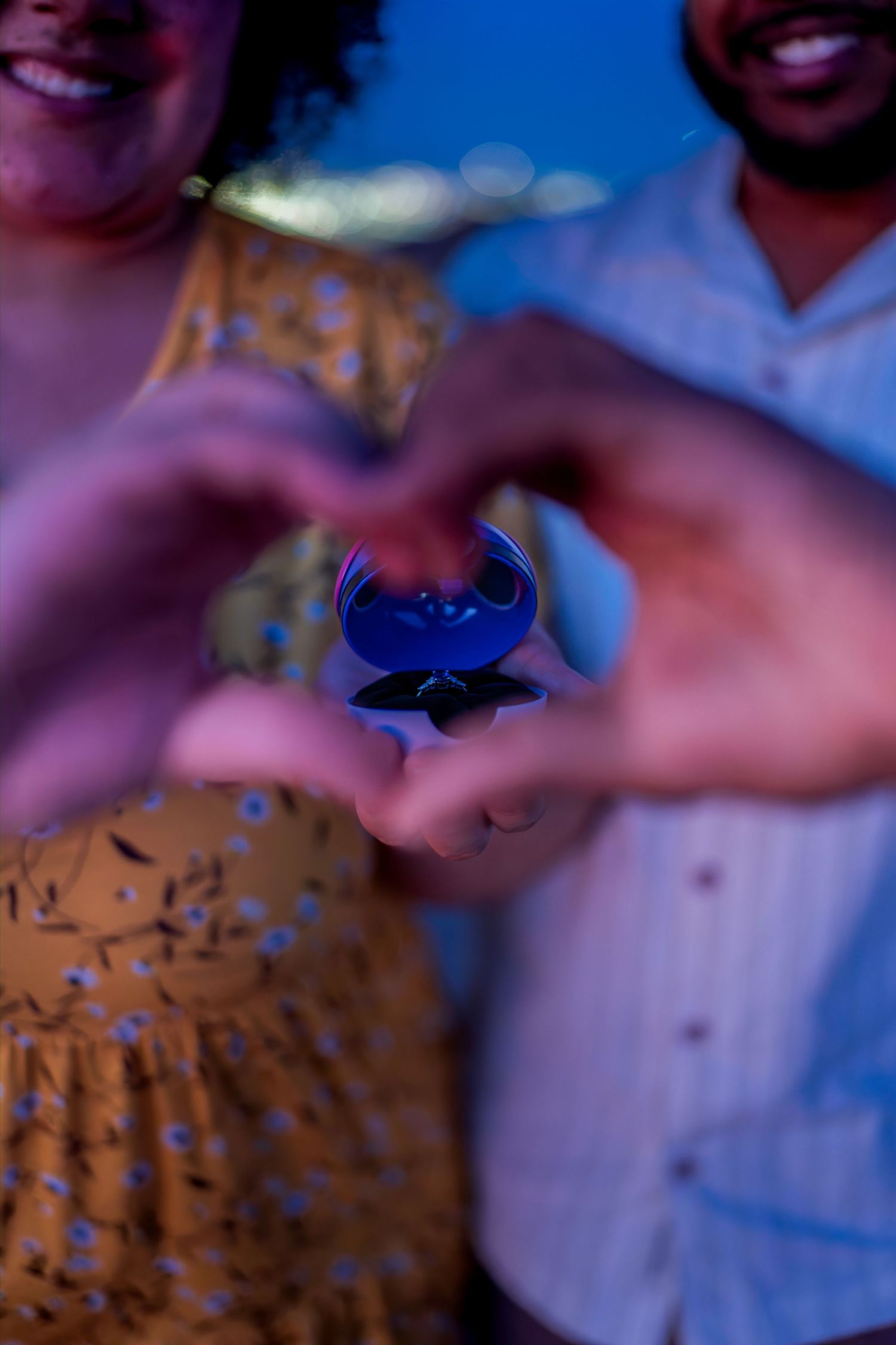 Couple making heart shape with hands around a blue ring box during romantic evening proposal.