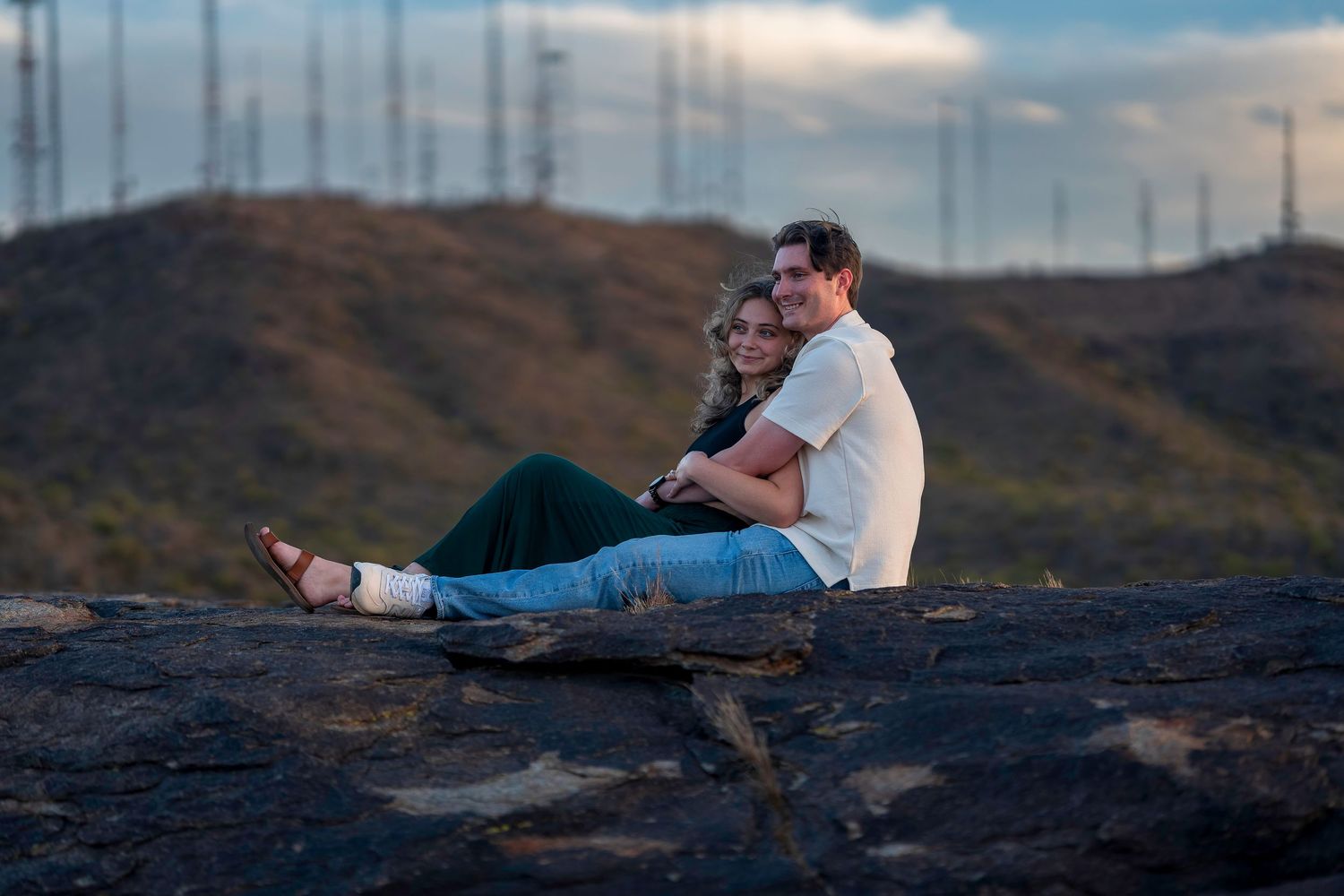 A couple sits closely together on a rock at sunset with communication towers in the background.