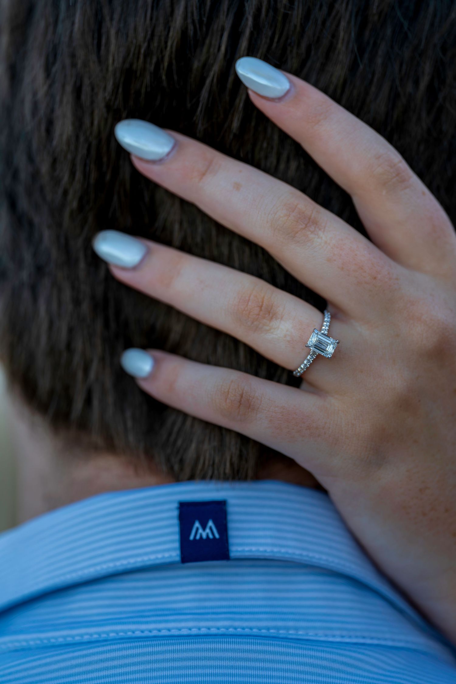 Close-up of an engagement ring on a woman's hand with white manicured nails resting on a man's neck.