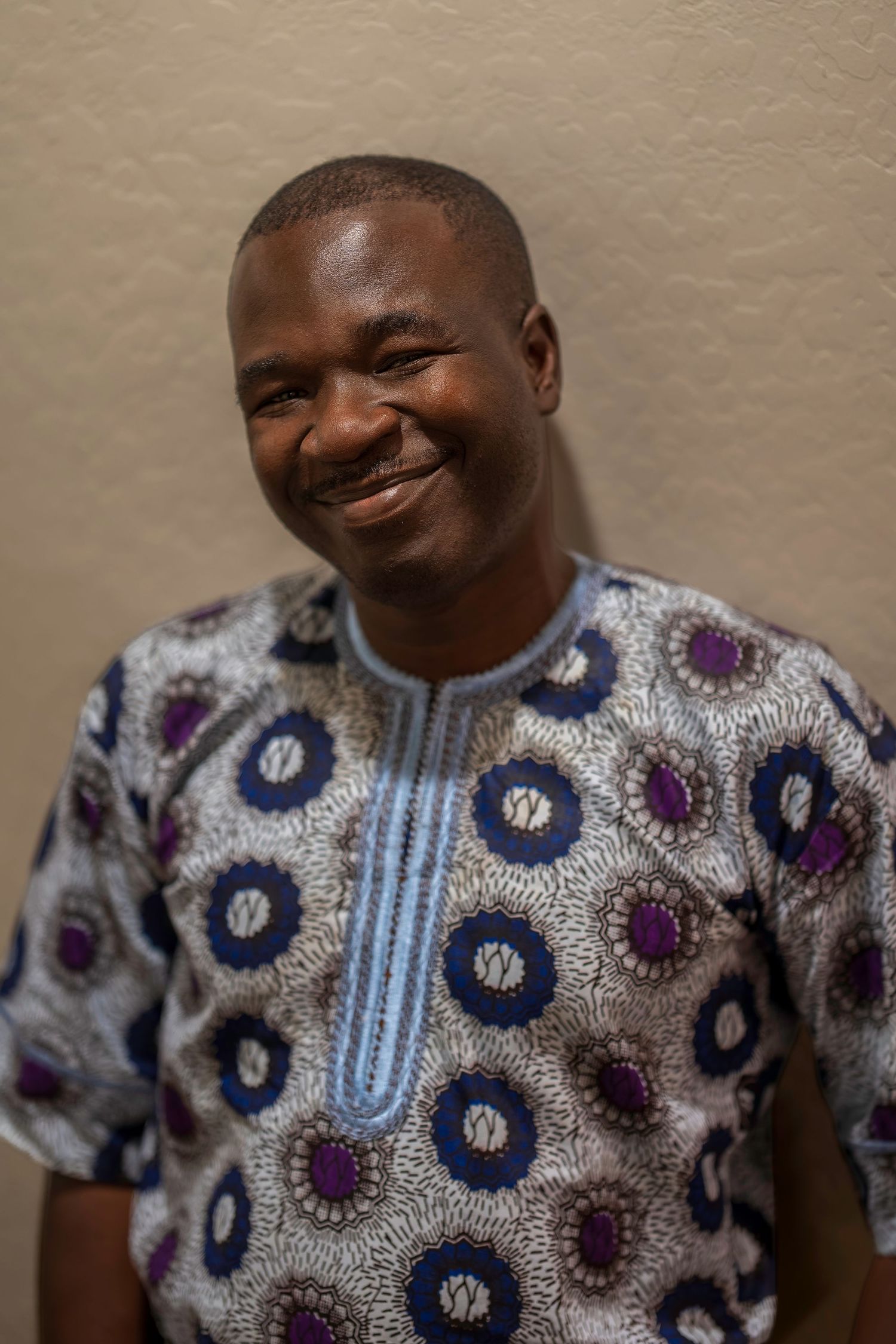 Smiling Black man in traditional African patterned shirt posing for a portrait against a beige background.