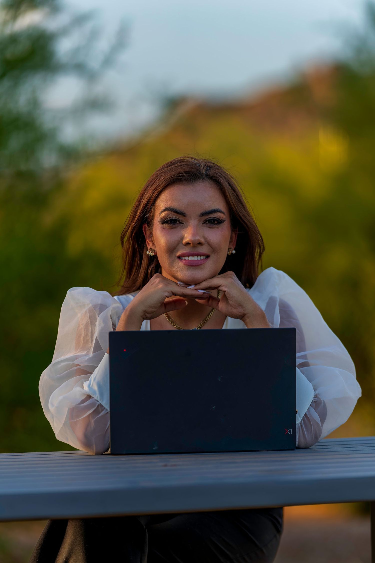 Smiling brunette woman in white sheer blouse leaning on laptop at outdoor table.