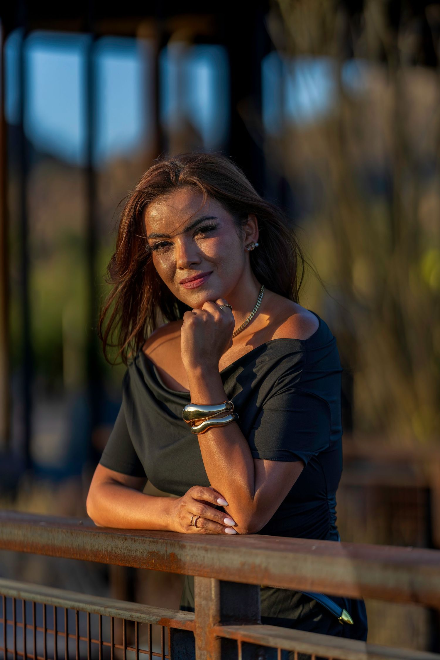 Young woman in black off-shoulder top leaning on rustic railing at golden hour.
