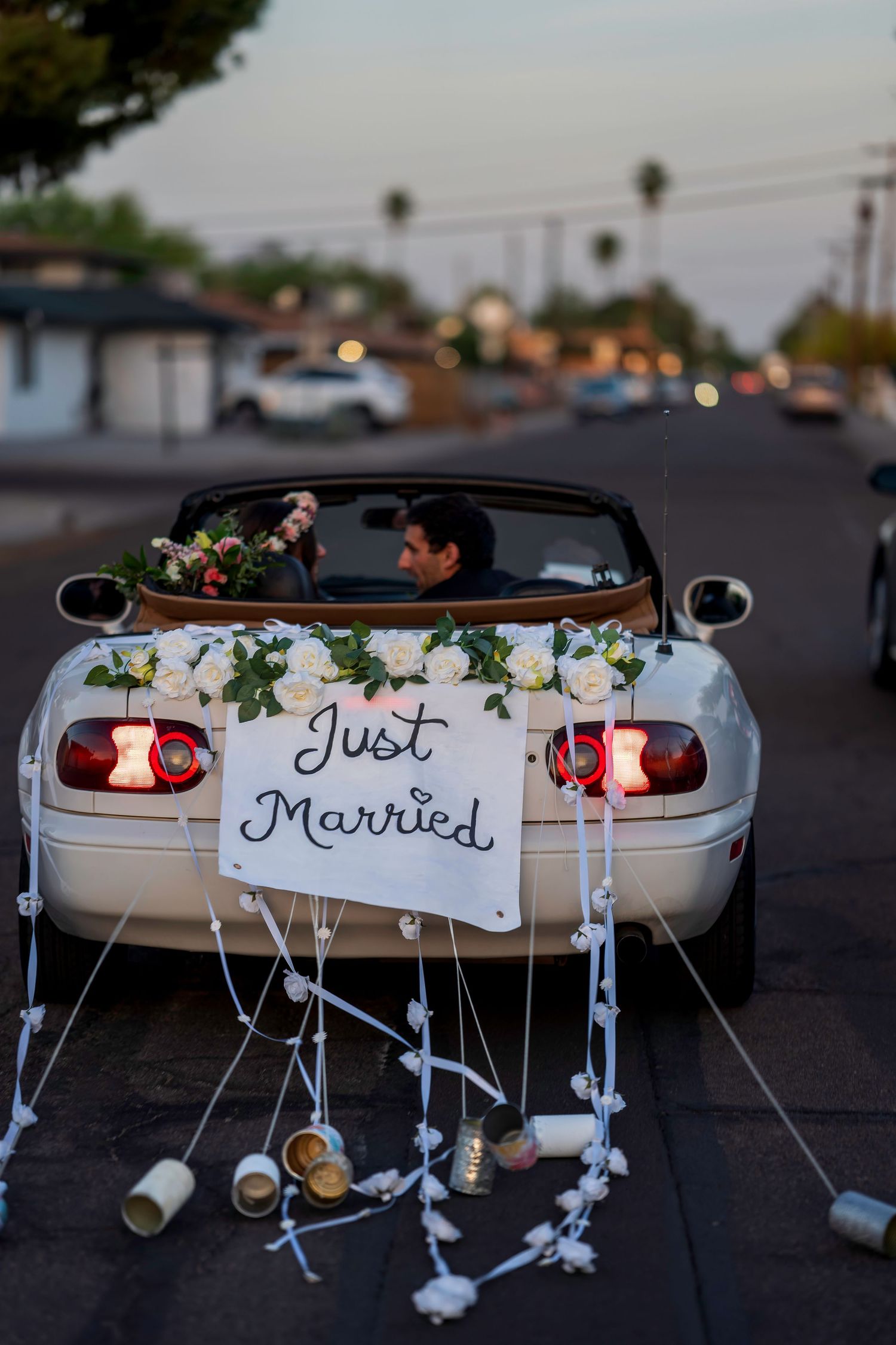 White convertible car decorated with roses and a Just Married sign with tin cans trailing behind on a suburban street.
