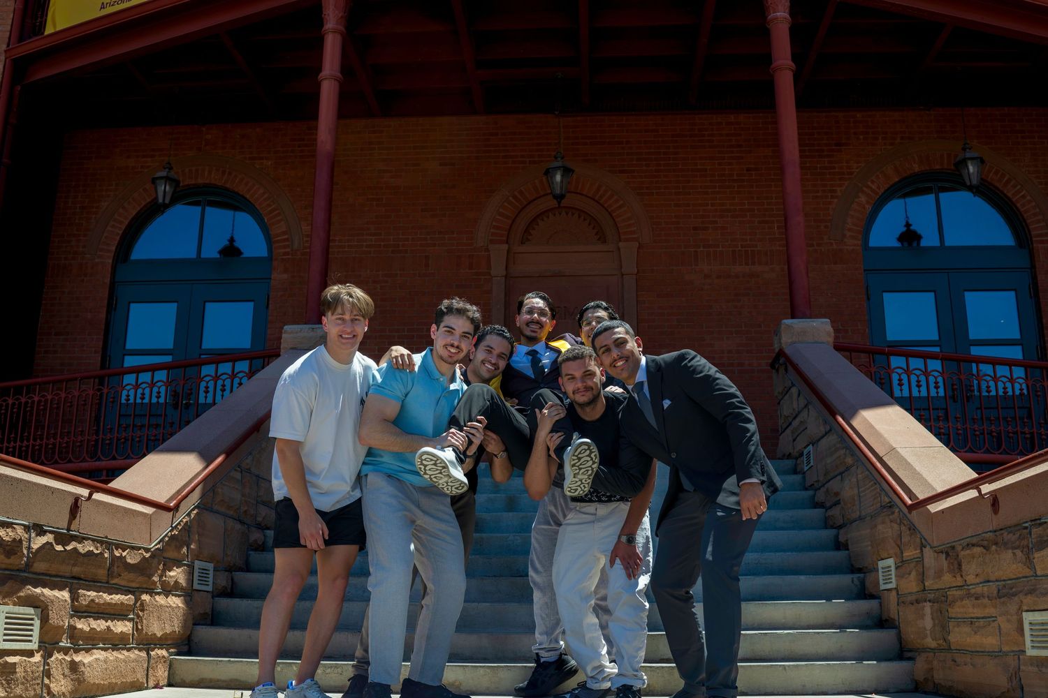 Group of young men posing on steps of historic red brick building on sunny day.