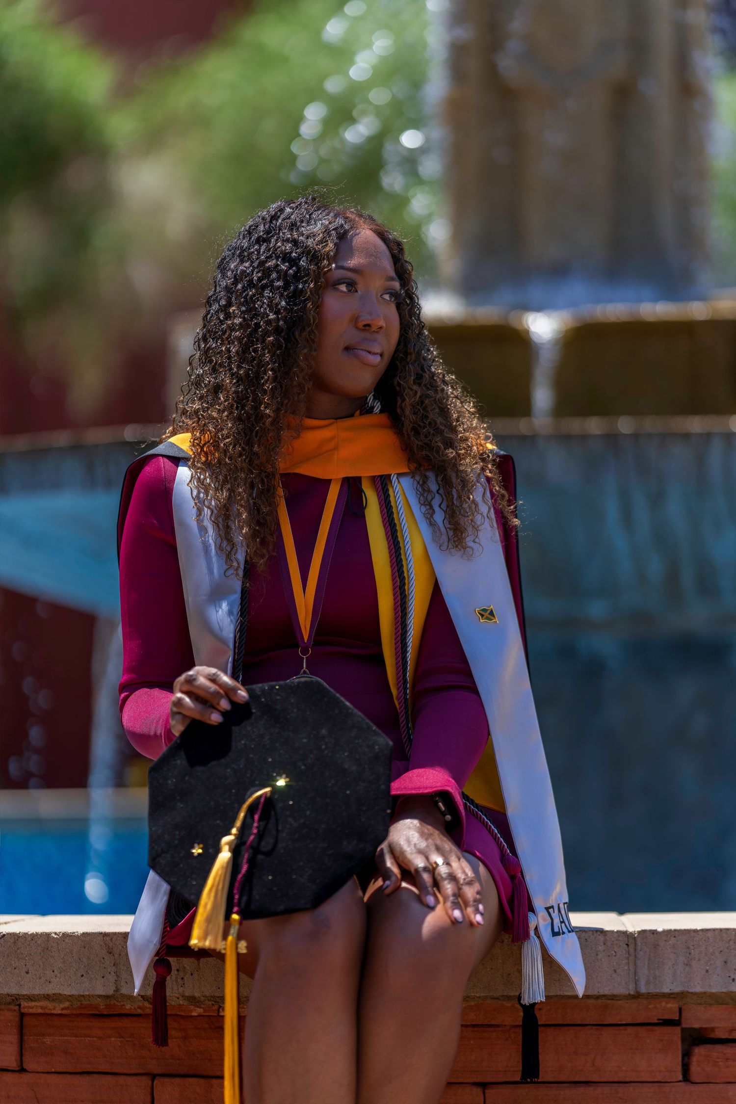 Graduate in maroon and white regalia holds cap while sitting by fountain on sunny college campus.