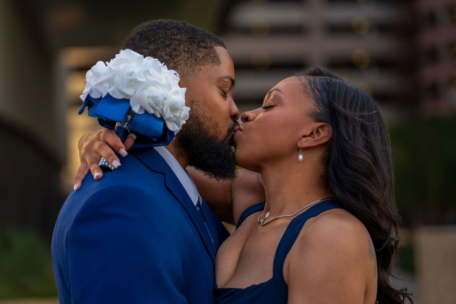 Couple sharing a kiss, groom in blue suit holding white bouquet, bride in navy dress at outdoor wedding.
