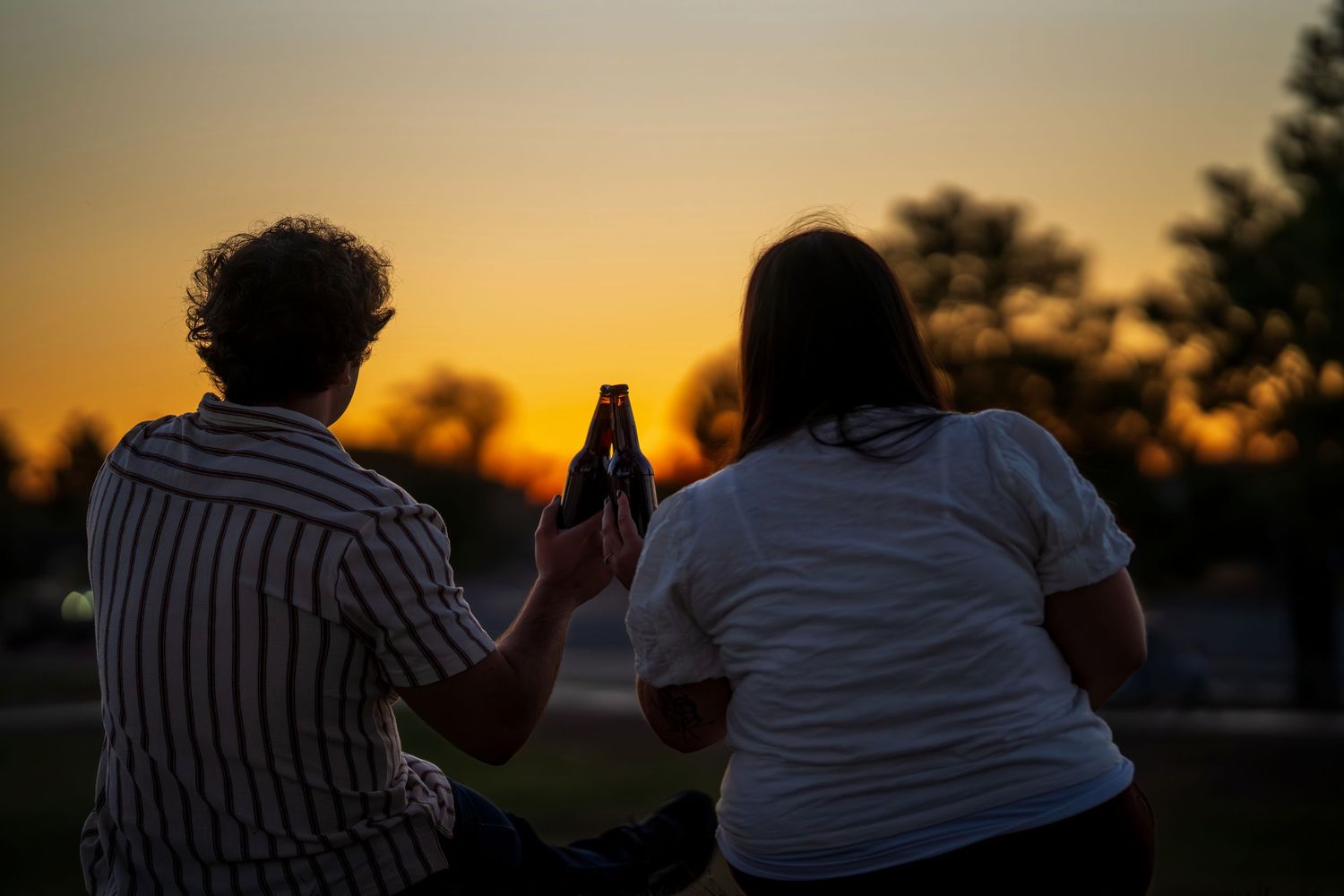 Two people sitting outdoors photographing a vibrant golden sunset with a smartphone at dusk.