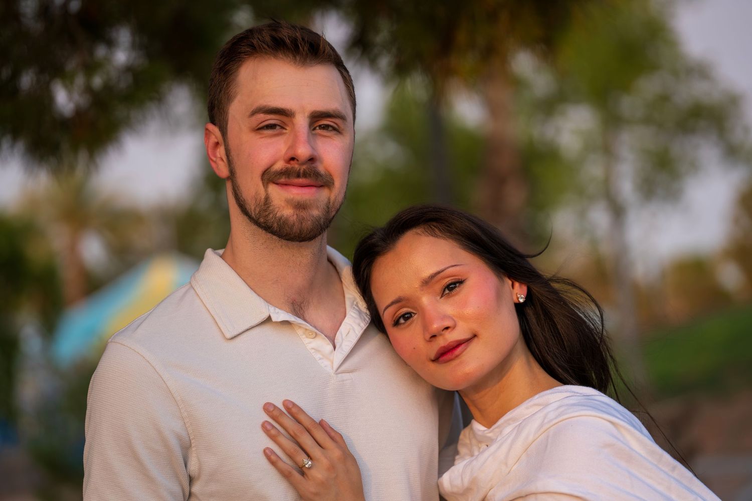 Smiling couple in white outfits posing outdoors with palm trees, woman displaying engagement ring.