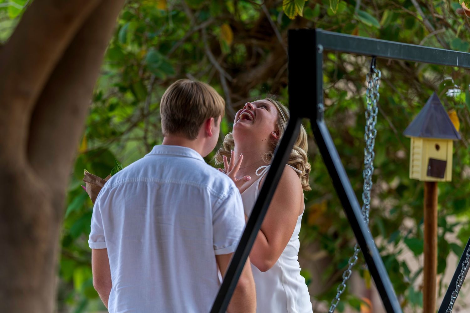 Couple laughing joyfully together outdoors near a swing set and birdhouse, surrounded by lush green trees.
