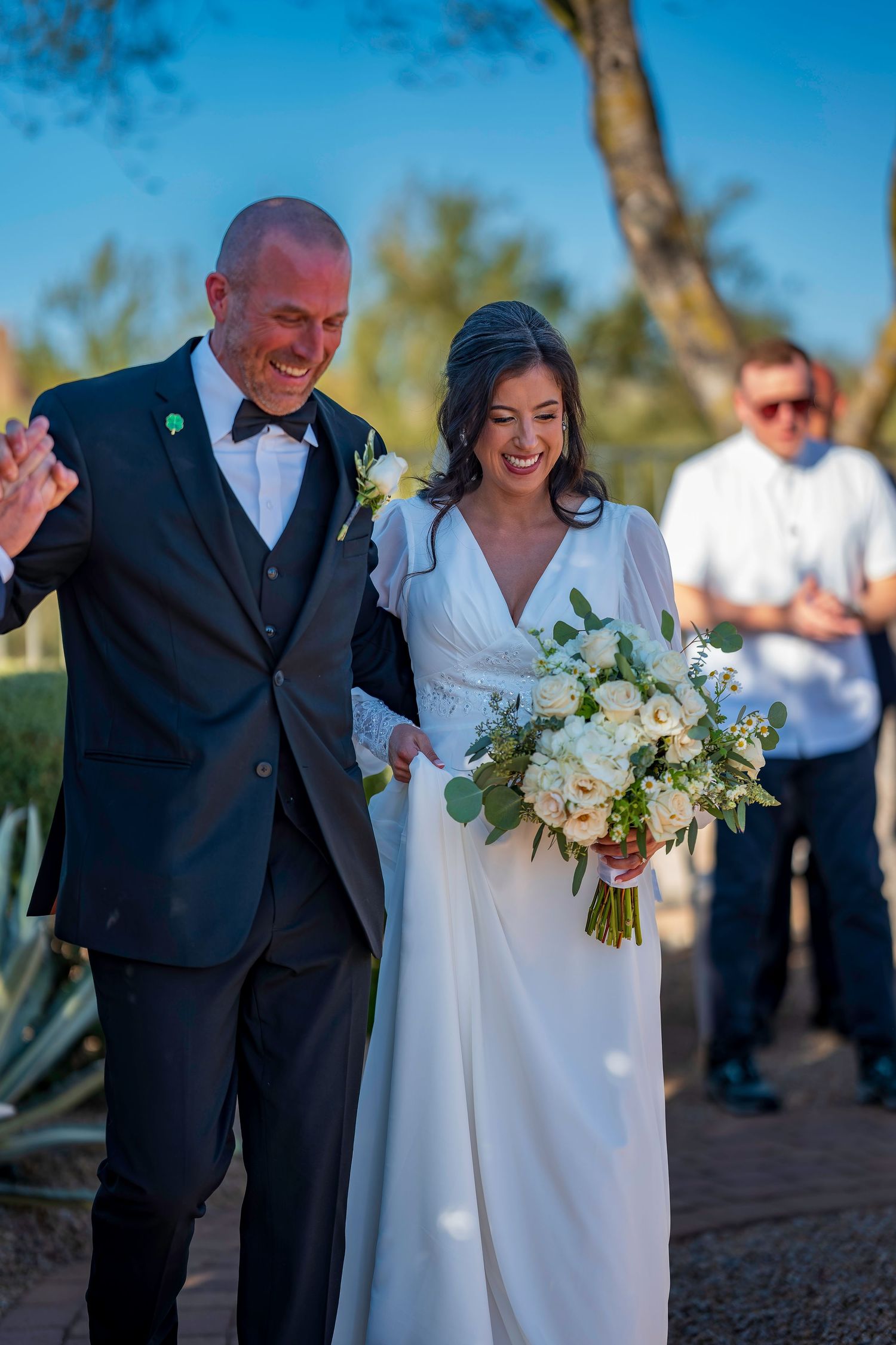 Newlyweds walk joyfully after outdoor wedding ceremony, bride holding white rose bouquet in sunny desert setting.