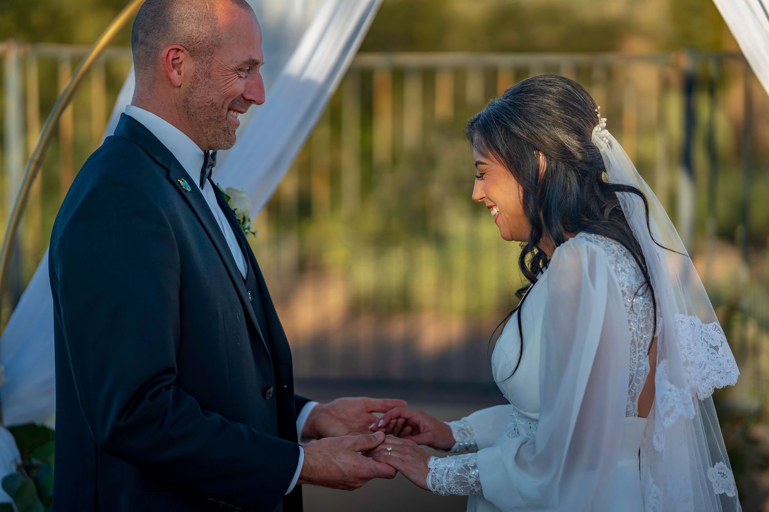 Groom smiles while placing ring on bride's finger during outdoor wedding ceremony at altar.