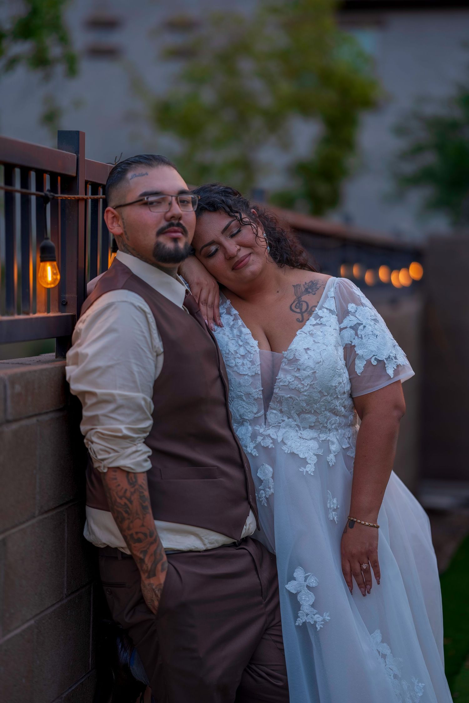 Bride rests head on groom's shoulder in romantic evening wedding portrait against brick wall with string lights.