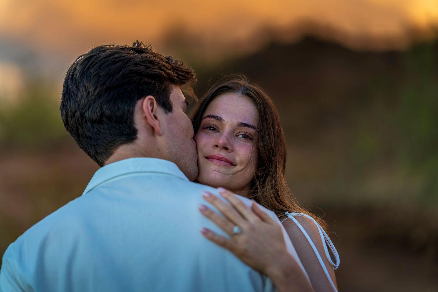A romantic couple embraces during golden hour sunset while wearing coordinating light blue and white outfits.