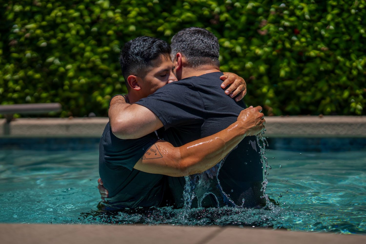 Two men embrace in a pool during a baptism ceremony, surrounded by lush green hedges.