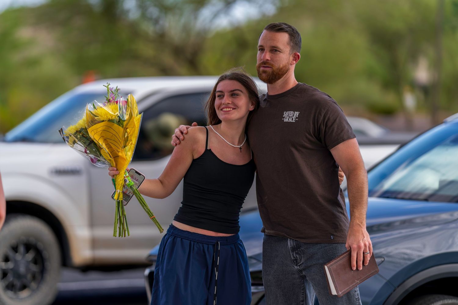 A couple stands together in a parking lot, the woman holding yellow flowers, both smiling warmly in evening light.