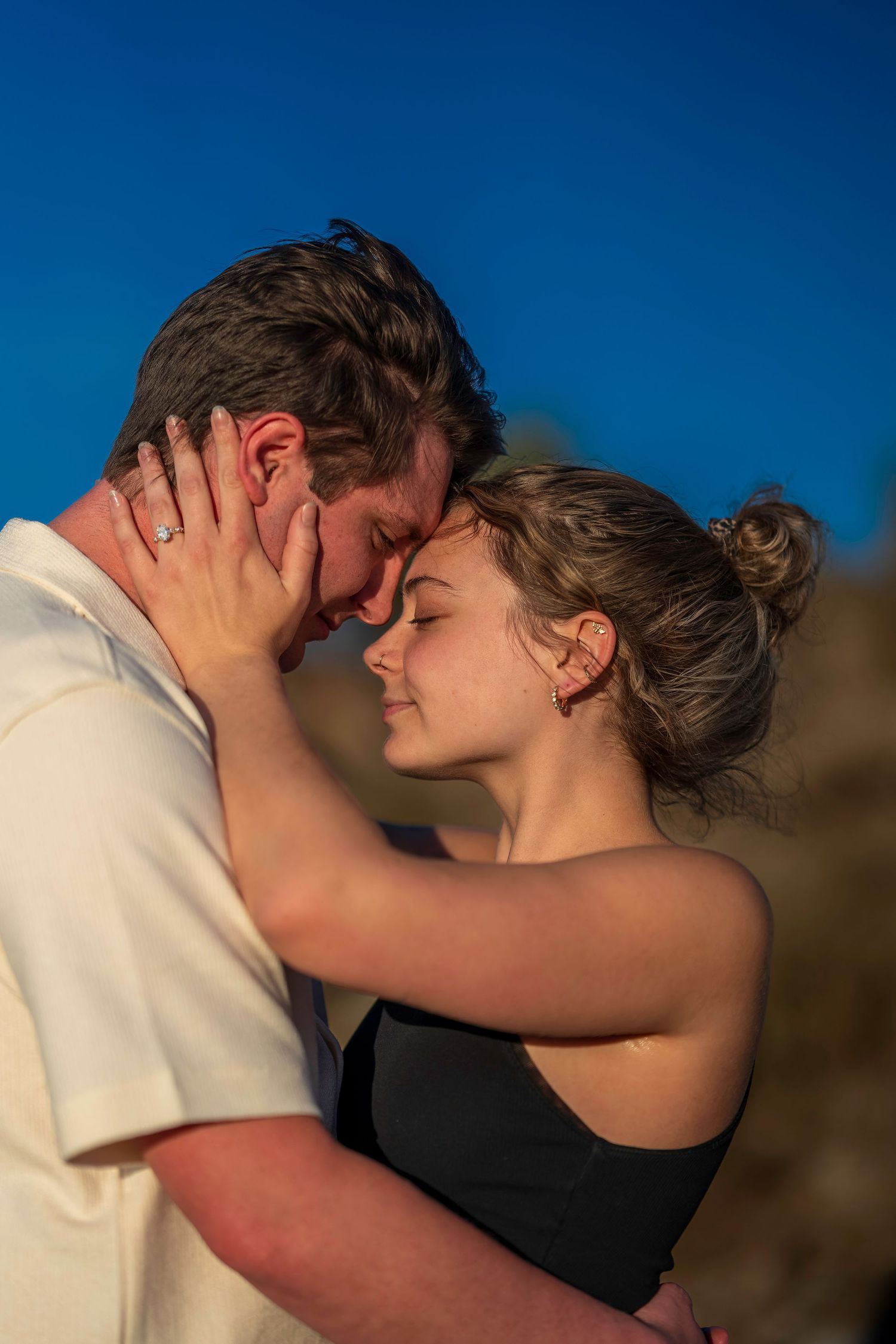 A young couple shares a romantic forehead-to-forehead embrace outdoors under a clear blue sky in golden hour light.
