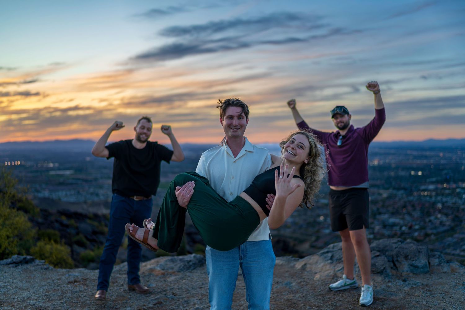 Group of four friends celebrating on a hilltop at dusk with a scenic city skyline in the background.