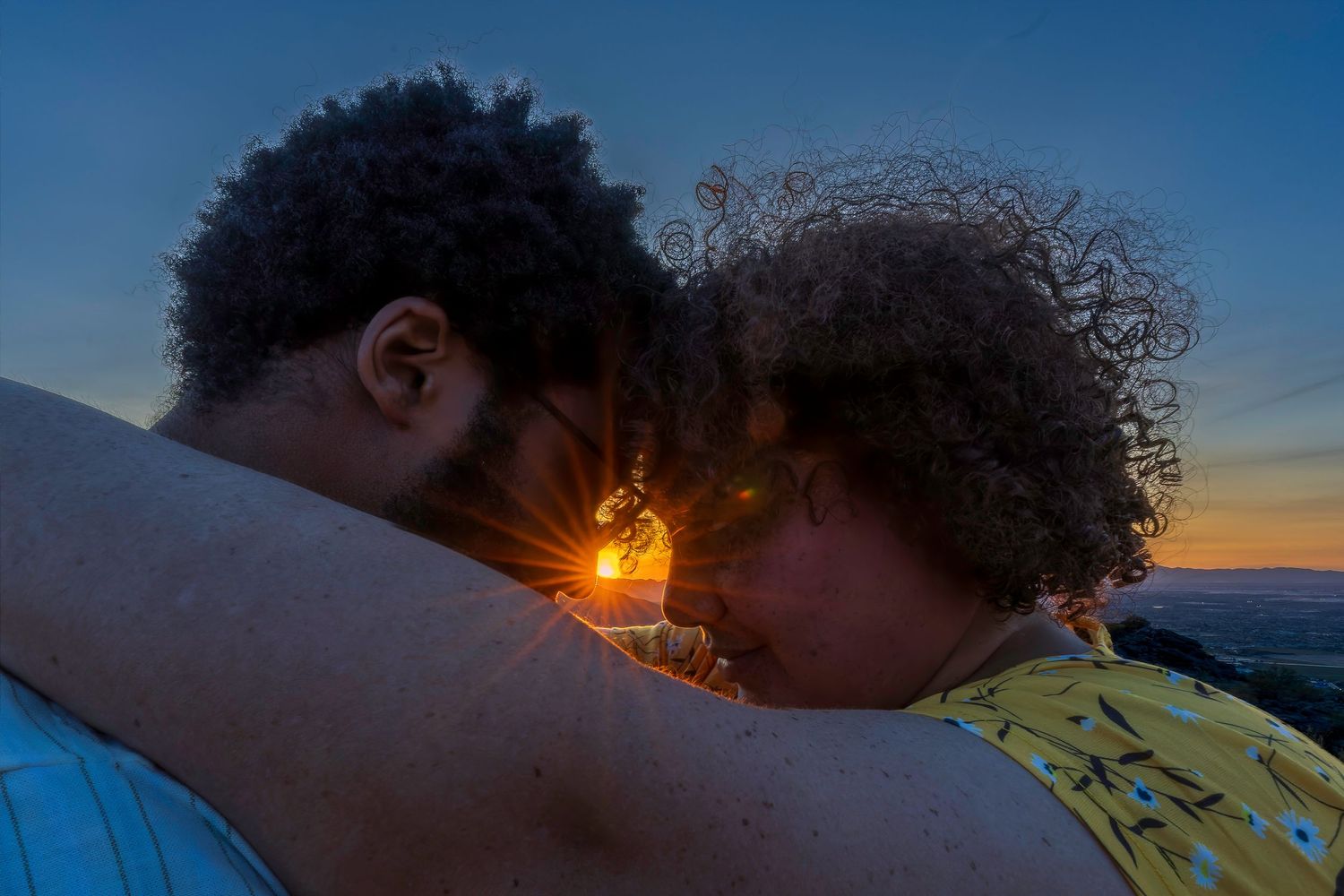 Two people with curly hair embrace forehead-to-forehead against a vibrant sunset sky.