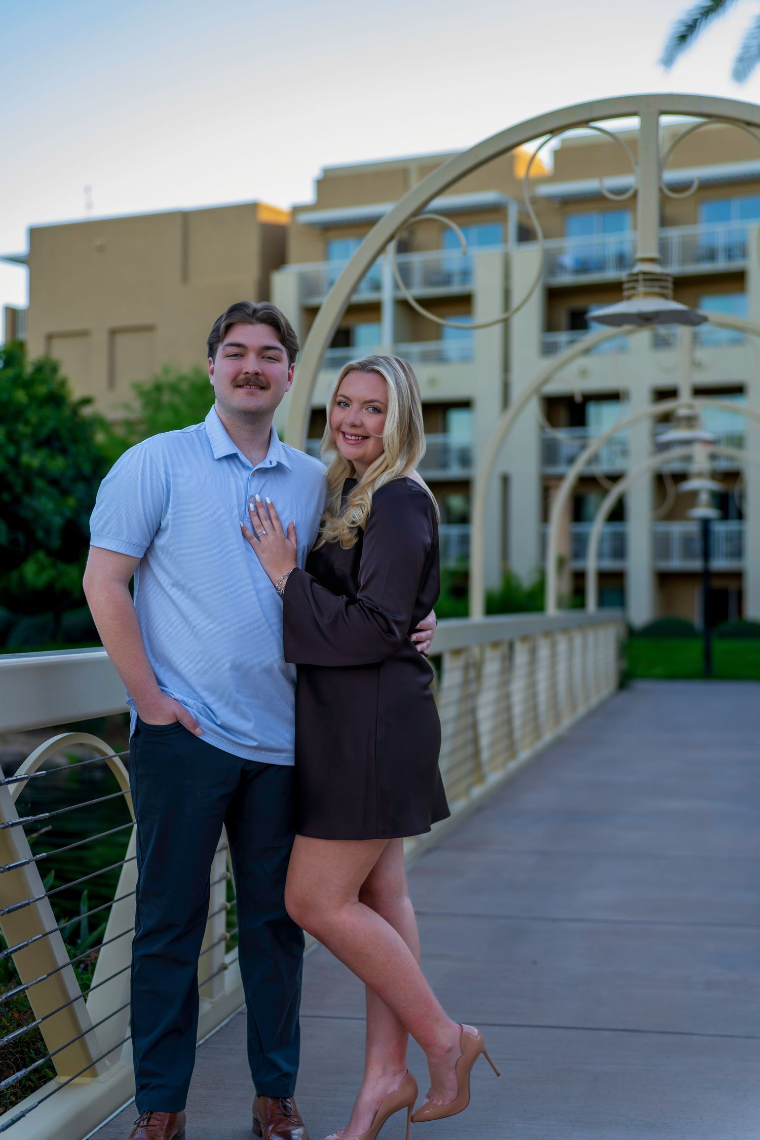 Engaged couple standing on decorative bridge at dusk, woman displaying diamond ring, urban backdrop.