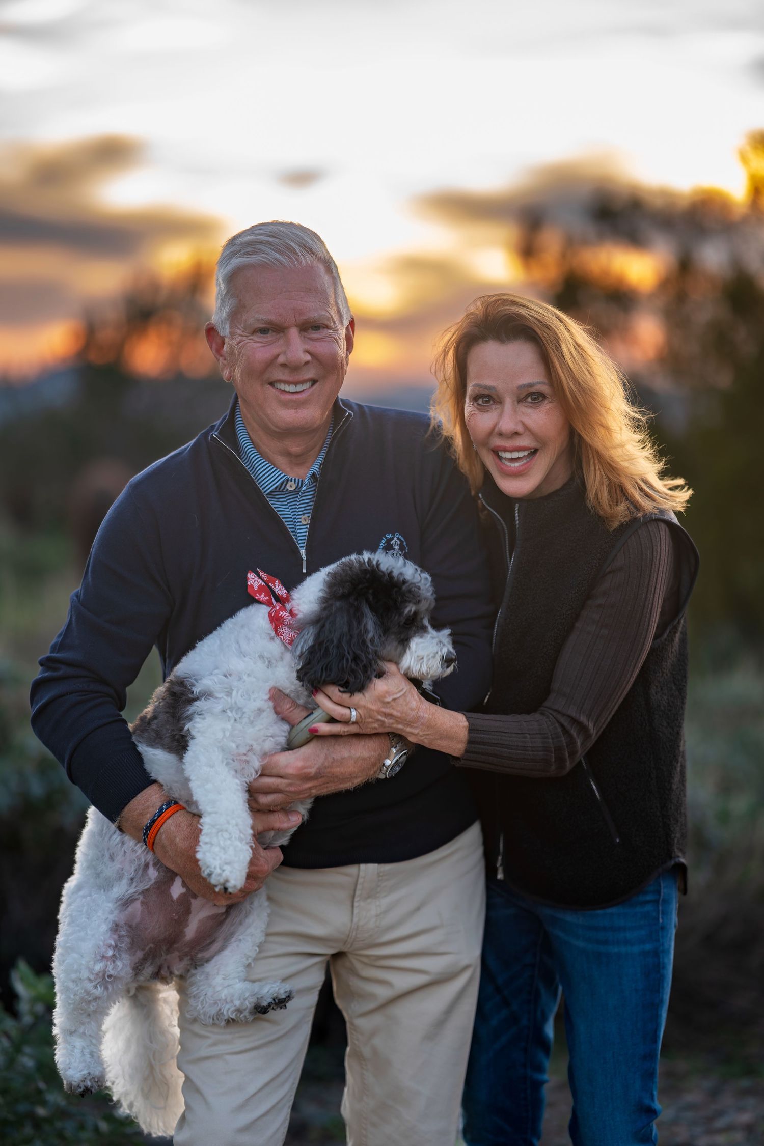 A couple holds a small black and white dog while posing outdoors during sunset.