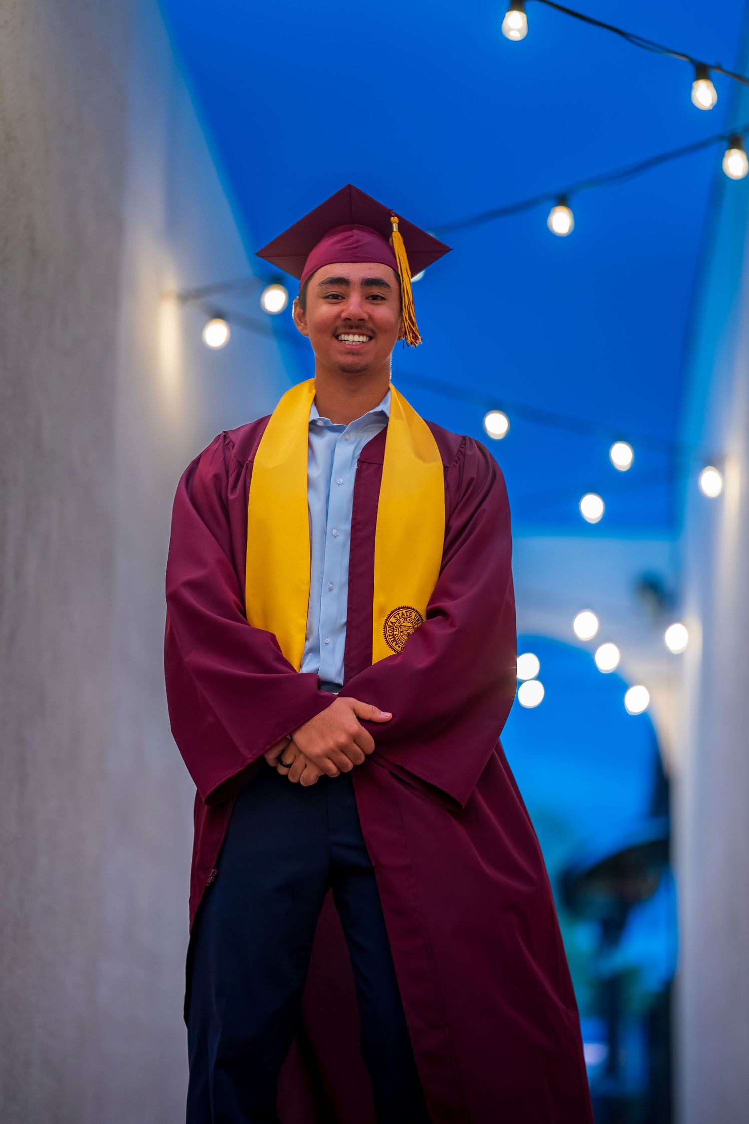 Graduate poses under string lights in maroon graduation gown and gold stole.