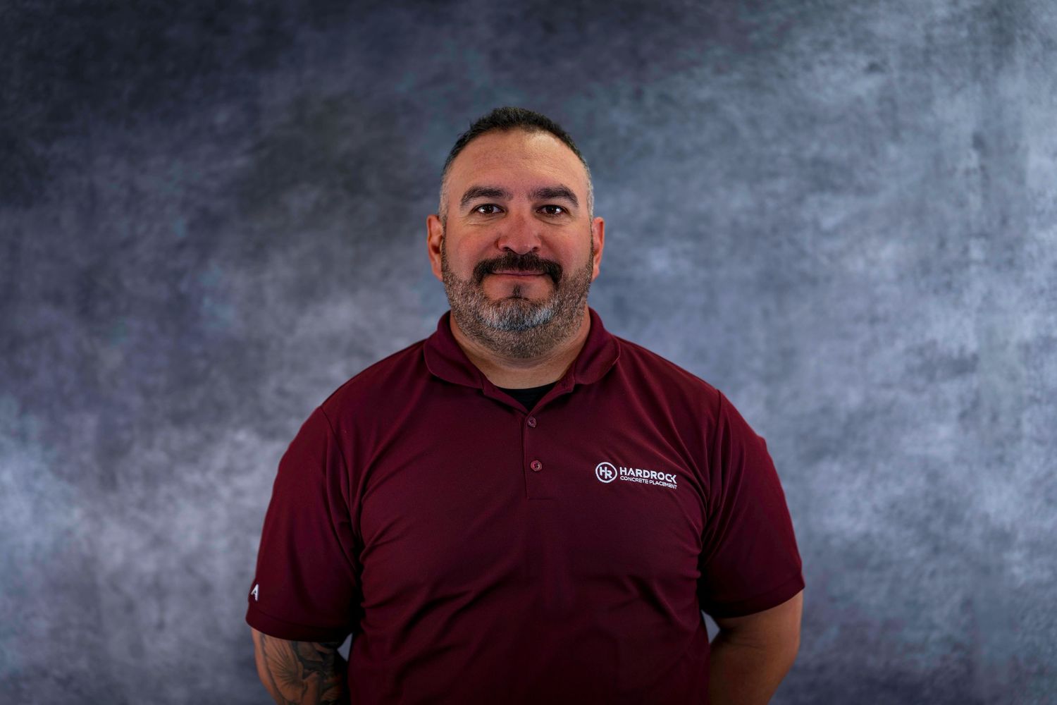 Employee wearing burgundy polo shirt with company logo against textured background.