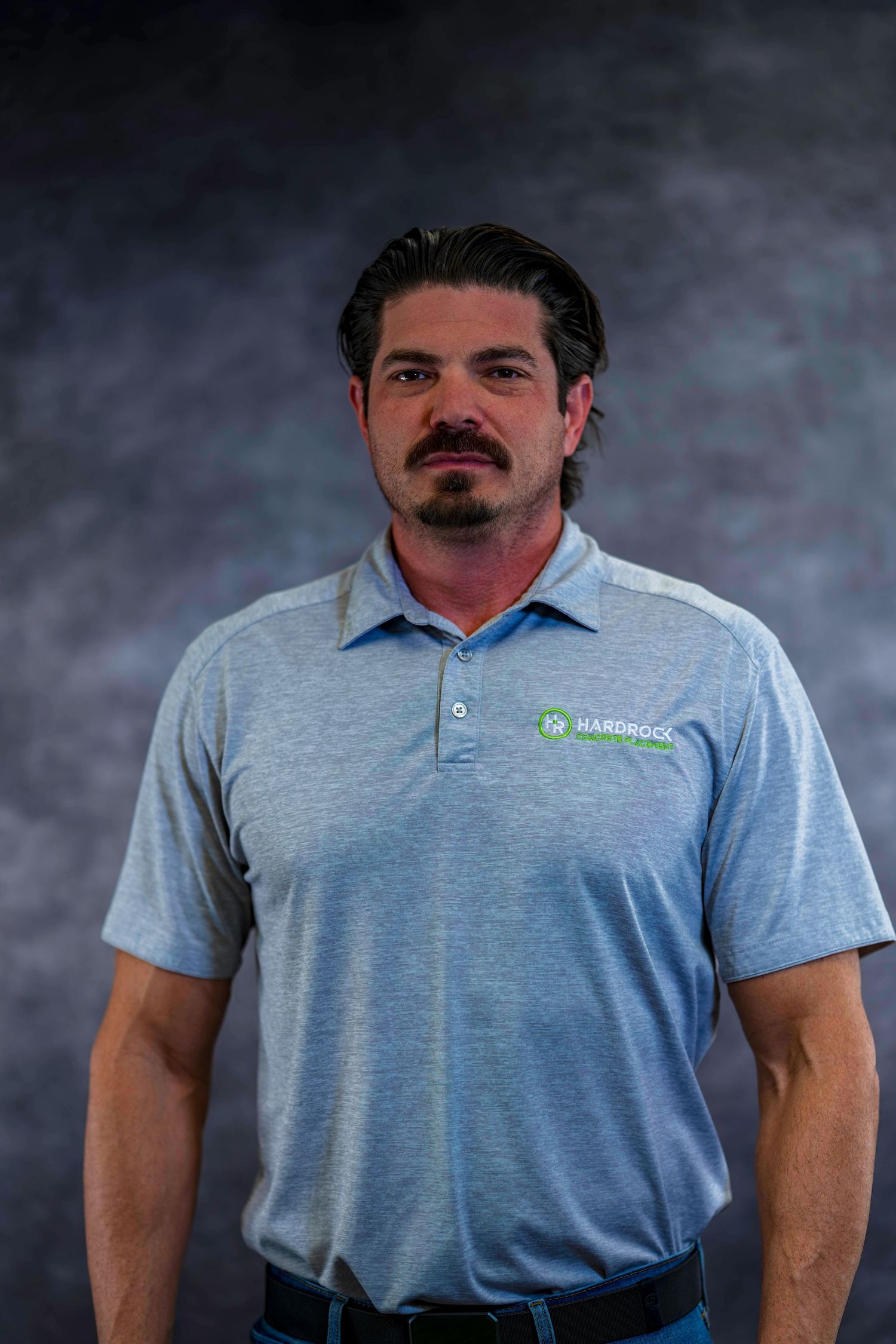 Professional headshot of employee in light gray polo shirt with company logo against dark background.