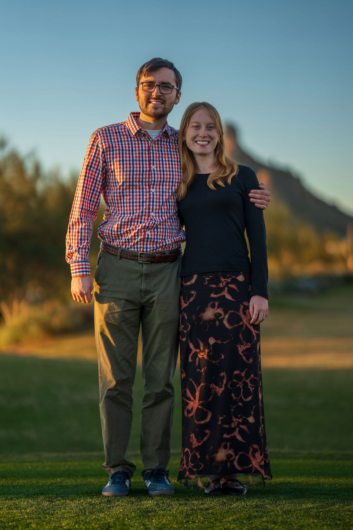 Couple posing together on grassy field with mountain silhouette in background at golden hour.