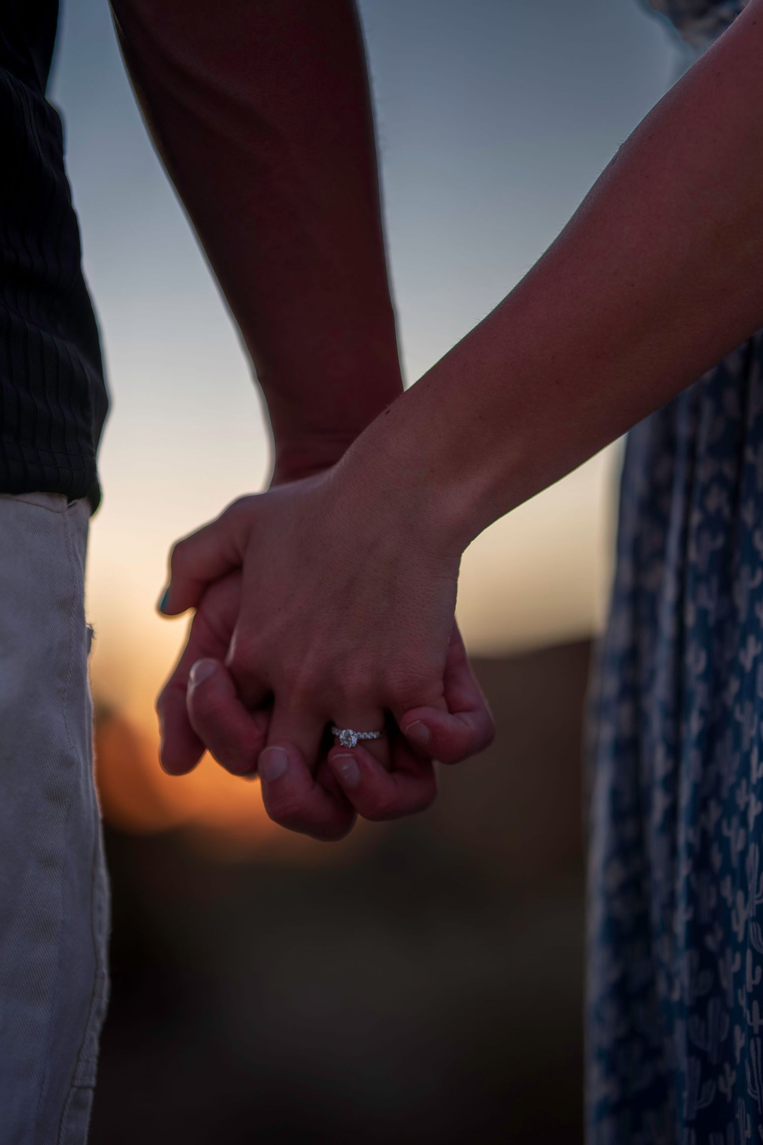Close-up of couple holding hands at sunset, woman wearing engagement ring.