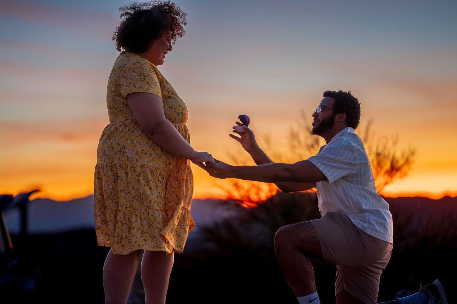 Man proposing with ring at sunset, woman in yellow floral dress, romantic outdoor engagement scene.