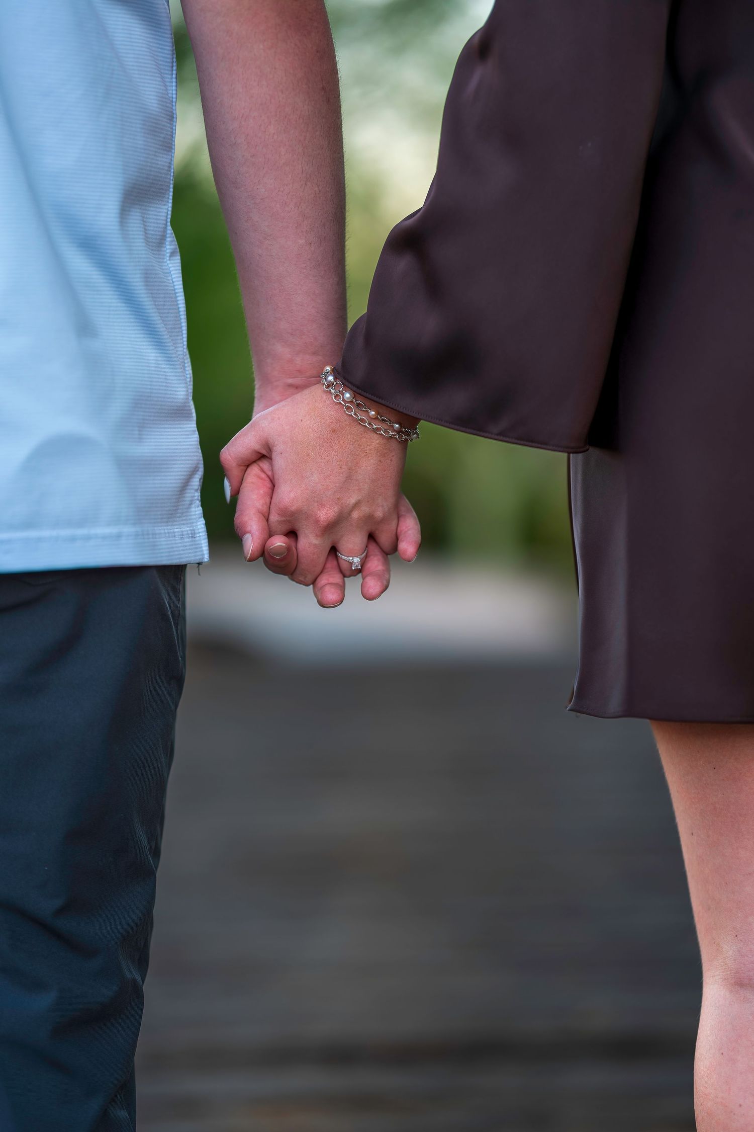 Couple holding hands gently outdoors, woman wearing a silver bracelet and engagement ring, in a park setting.