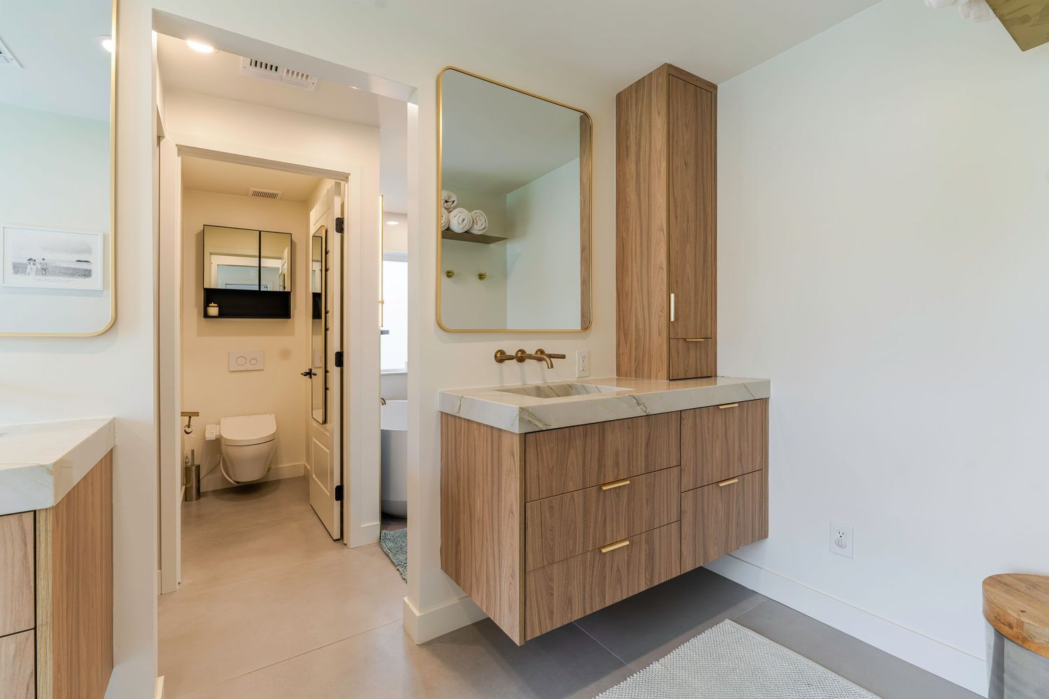 Modern bathroom vanity with floating wood cabinet, gold fixtures, framed mirror, tall storage tower, and view to toilet.