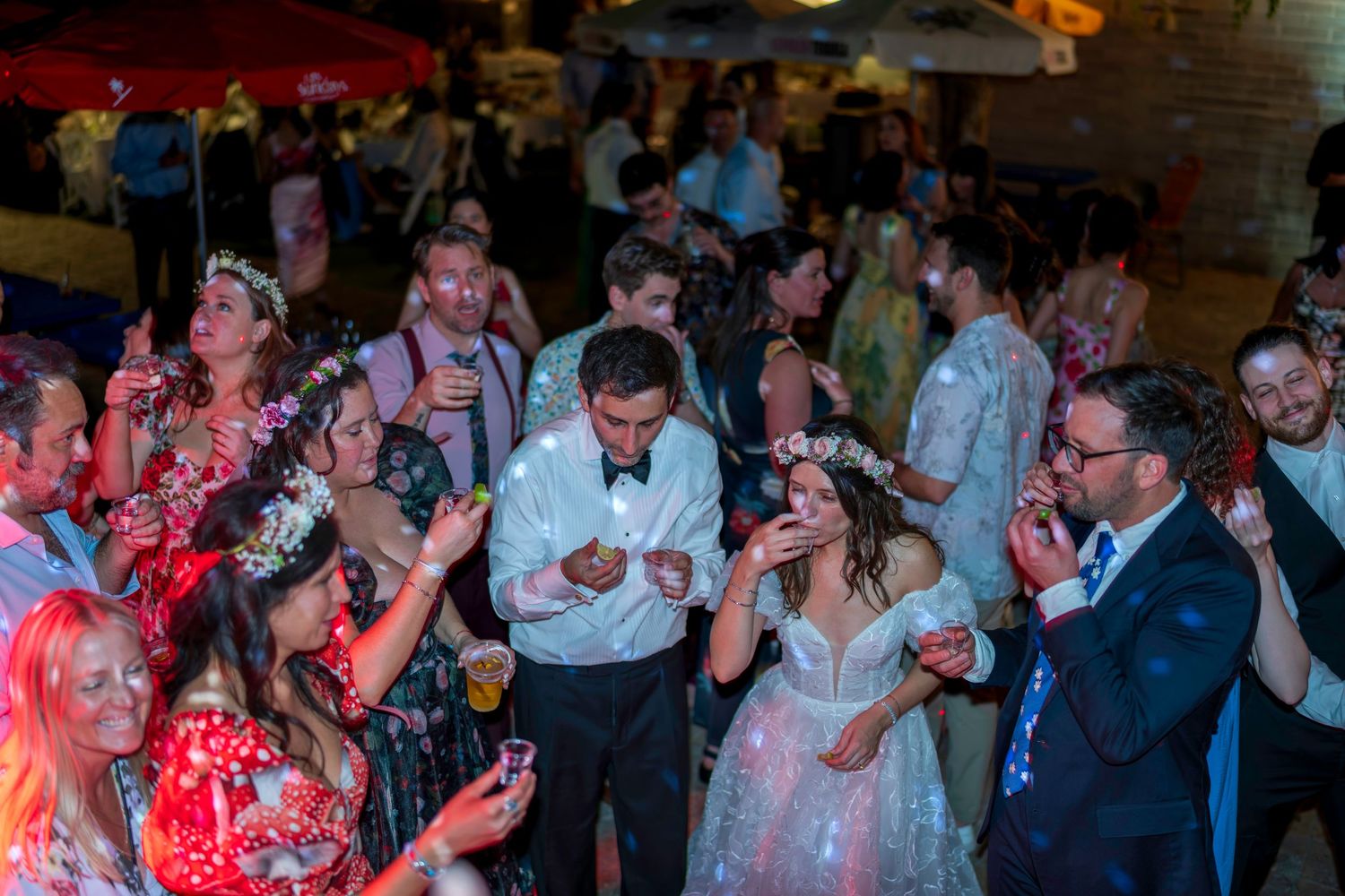 Bride and groom dancing joyfully with guests at a lively wedding reception party under colorful lights.