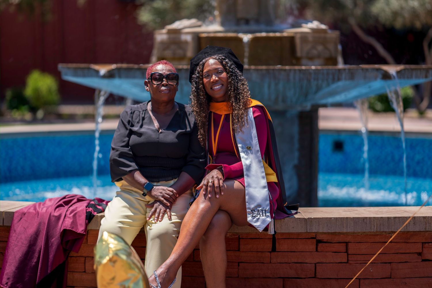 Two women smile together by a fountain, one wearing a maroon and gold graduation stole.