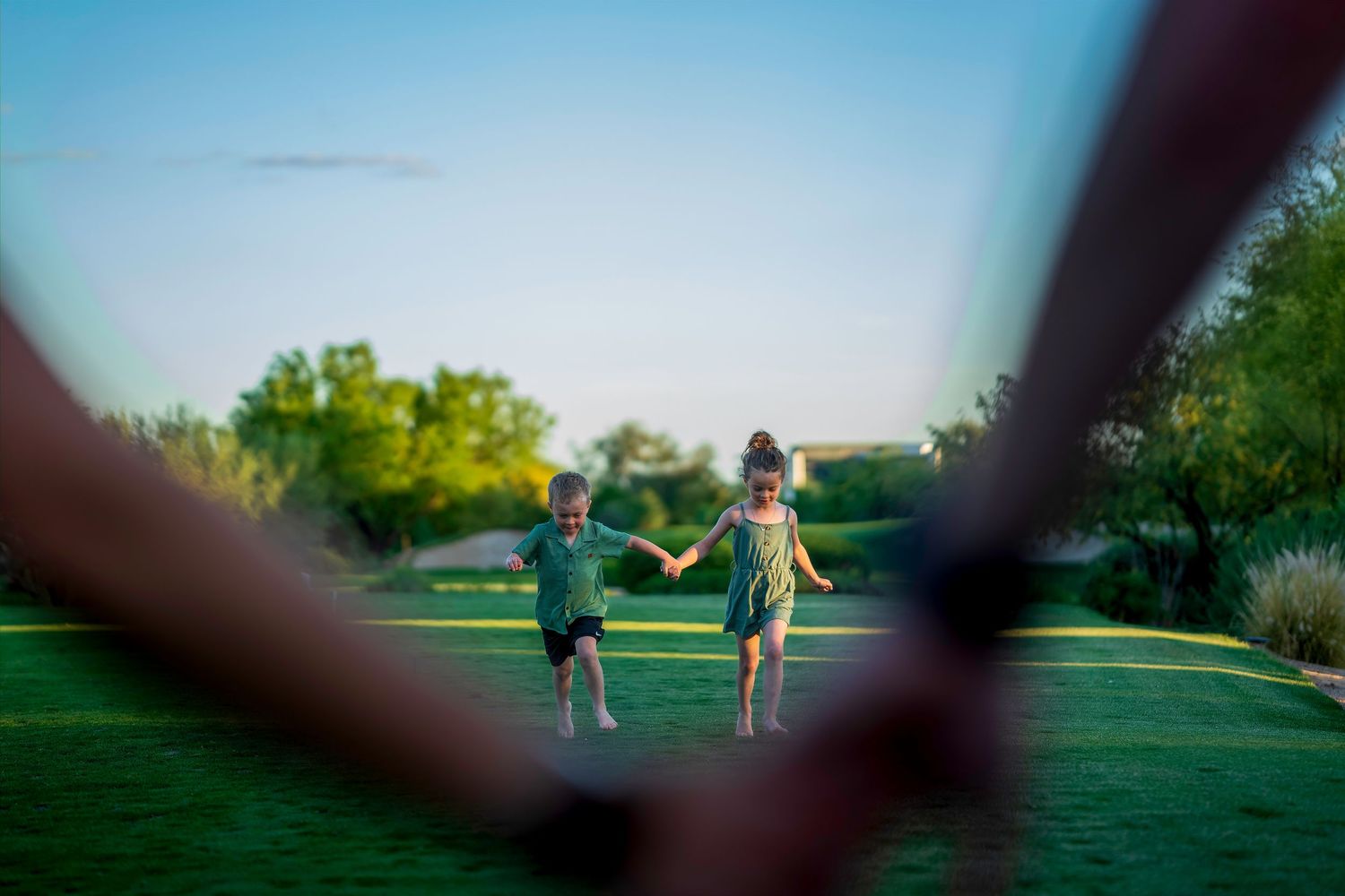 Two young children holding hands running across a lush green lawn toward the camera on a sunny day.