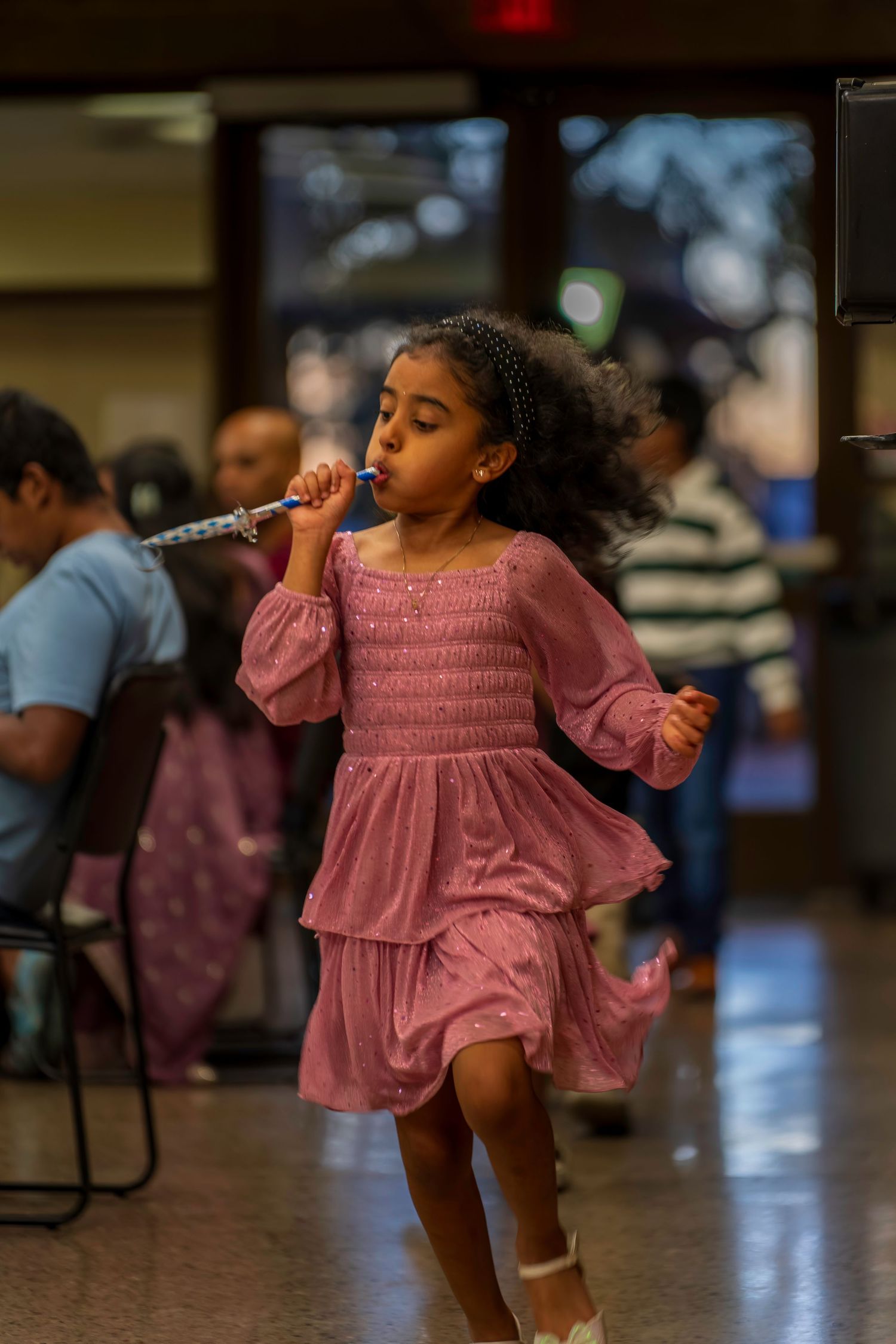 Young girl in pink dress dancing and blowing a party horn at an indoor celebration event.