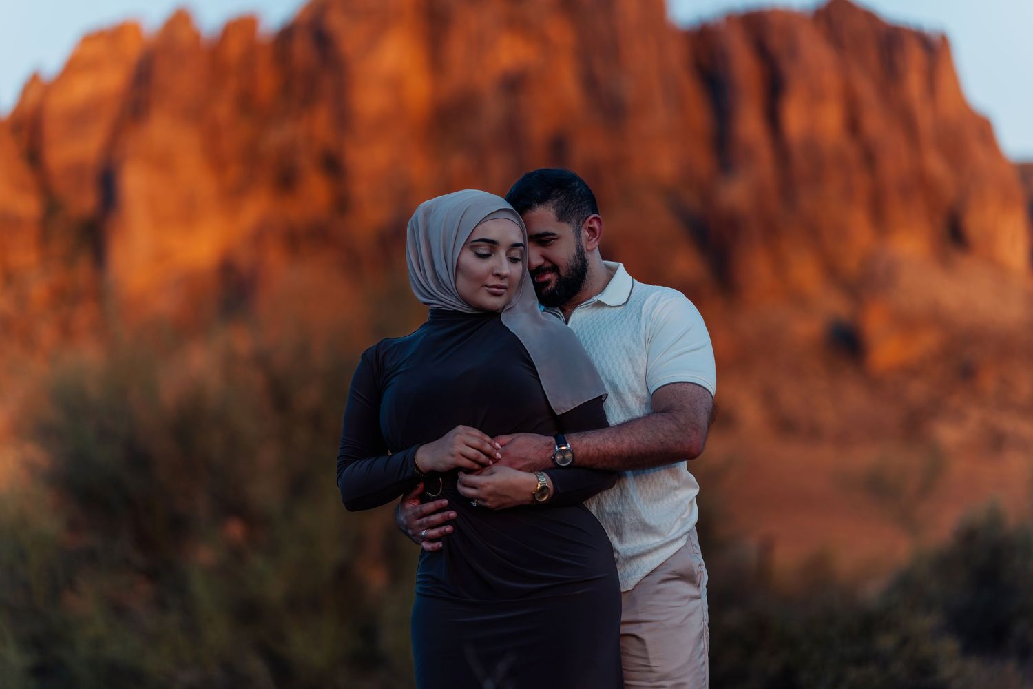 Couple embracing in front of dramatic red rock mountains at sunset in desert landscape.