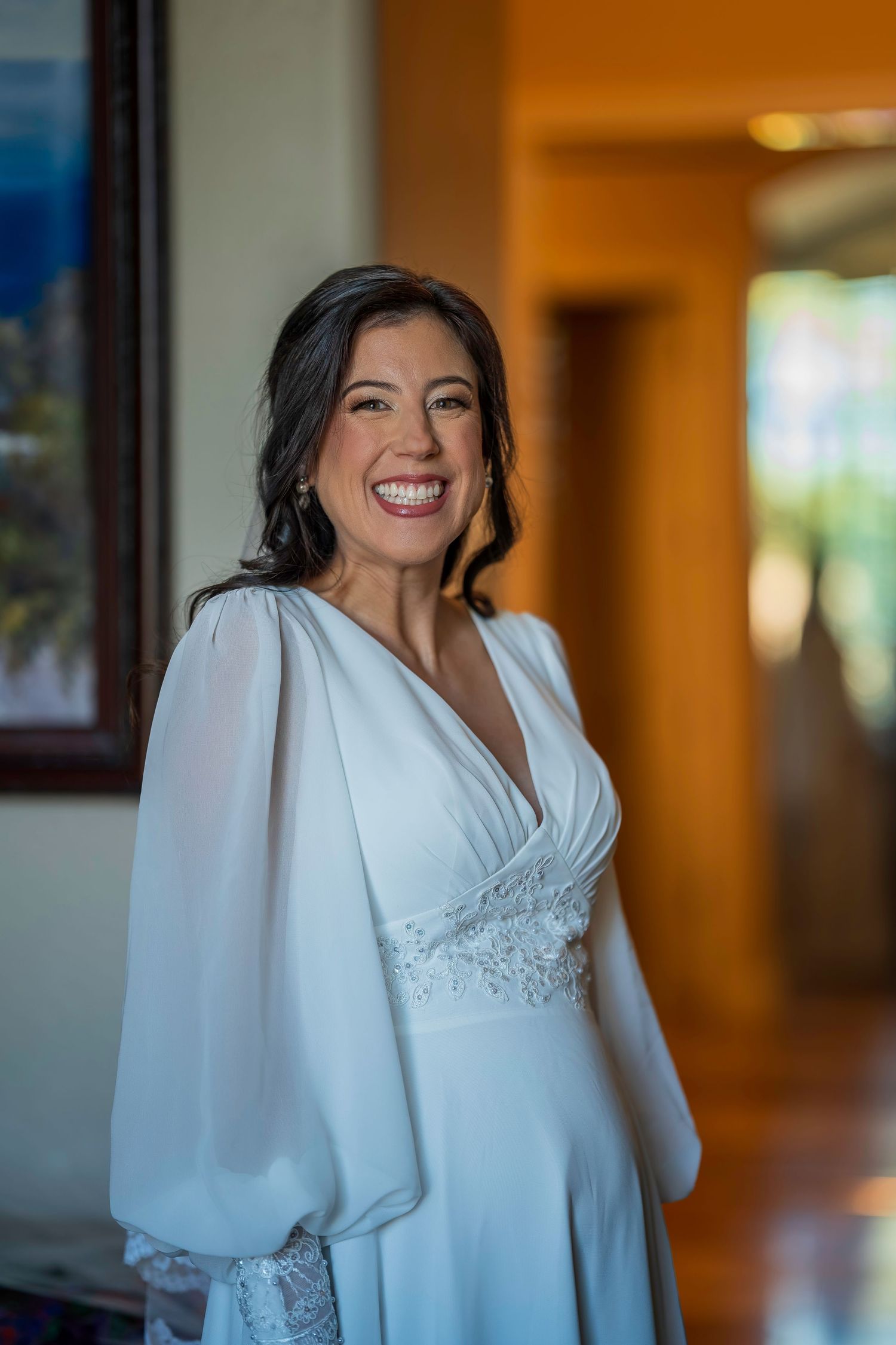 Smiling bride in elegant white long-sleeve wedding gown with lace detail posing indoors before her wedding ceremony.