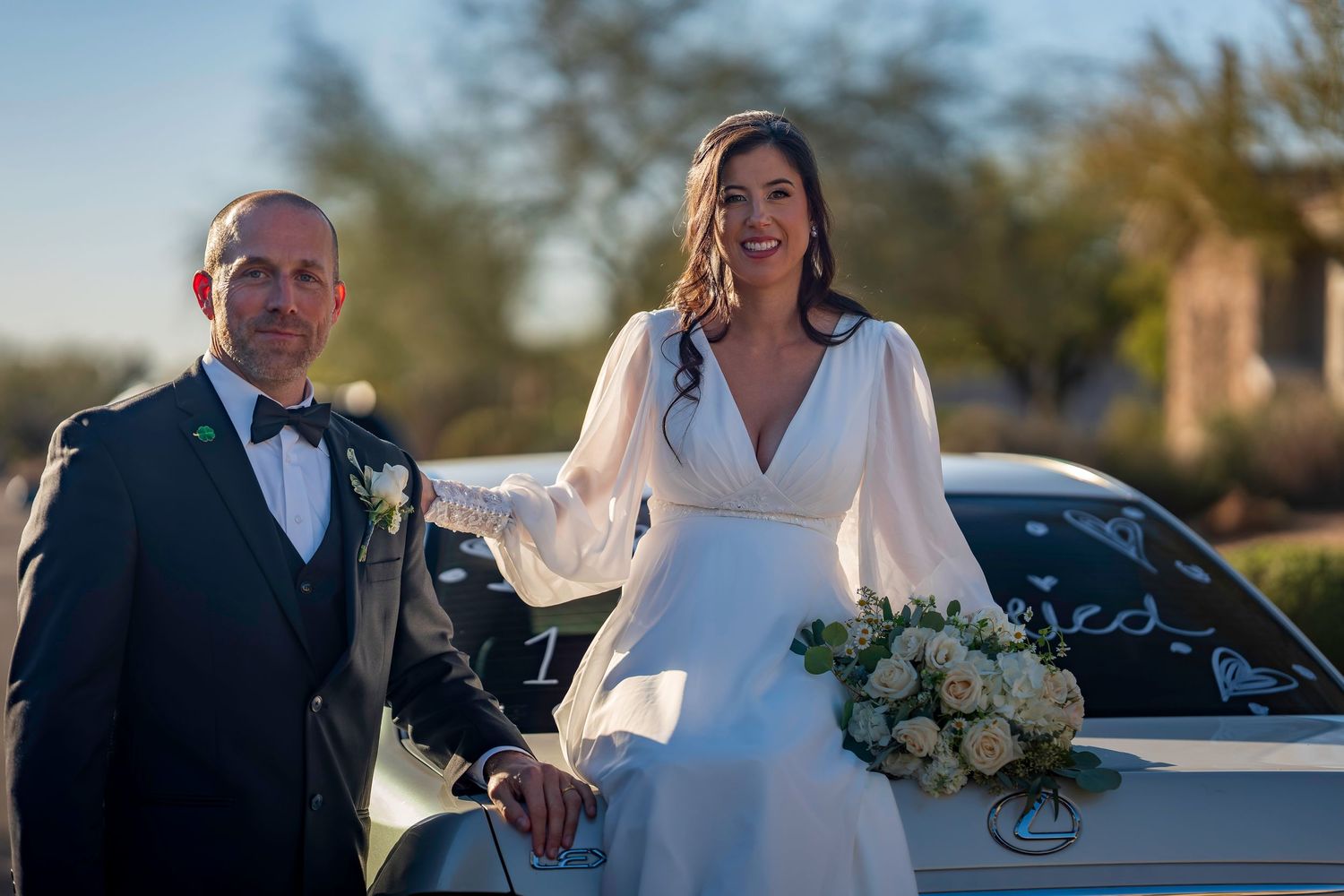 Bride and groom smile together leaning on a decorated car at outdoor wedding celebration.