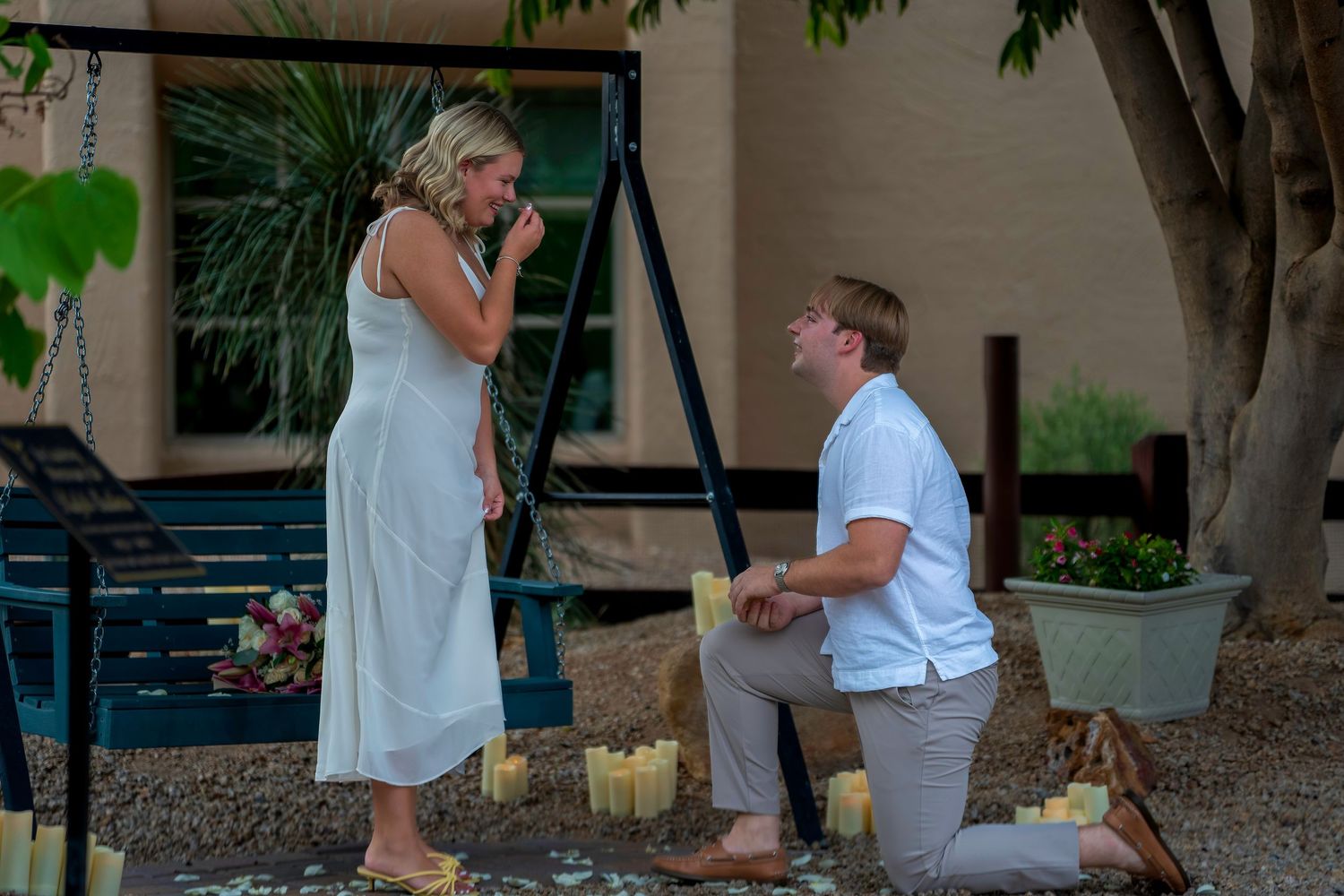 Man proposing on one knee to woman in white dress surrounded by candles outdoors.