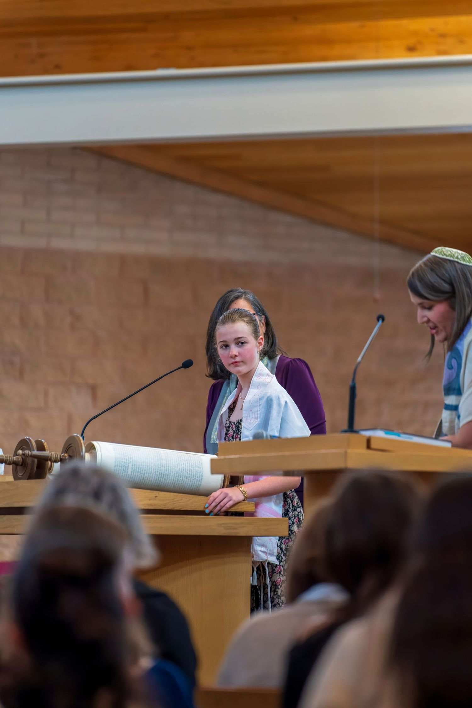 Young girl reads from Torah scroll at bat mitzvah ceremony with woman beside her at synagogue podium.